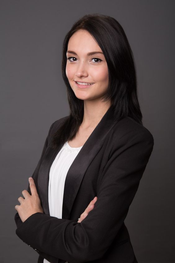 Young professional woman in black blazer and white blouse smiling with arms crossed against gray background.