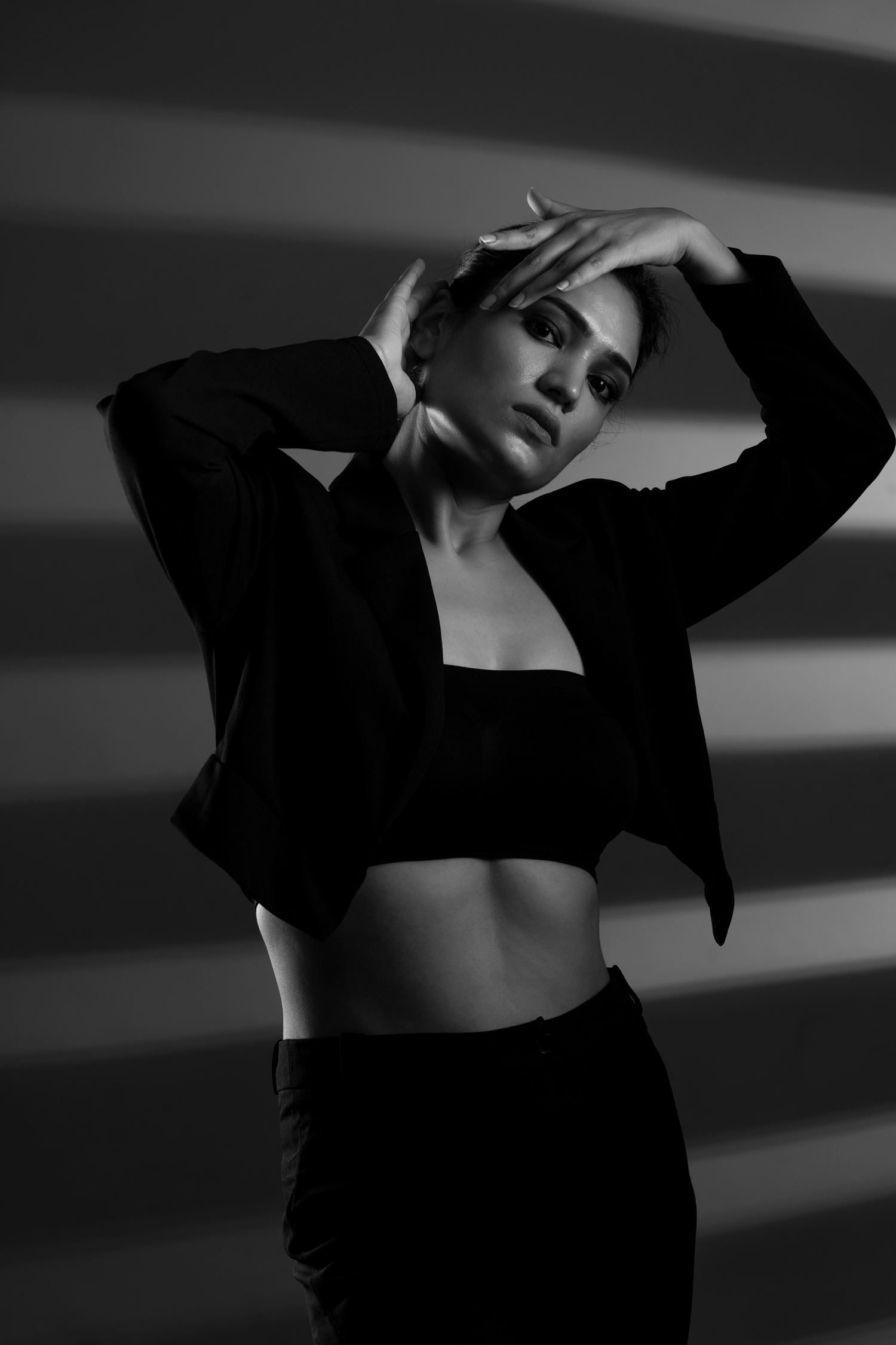 Black and white photo of a woman in a crop top and blazer posing with hands raised near her head against a striped shadow wall.