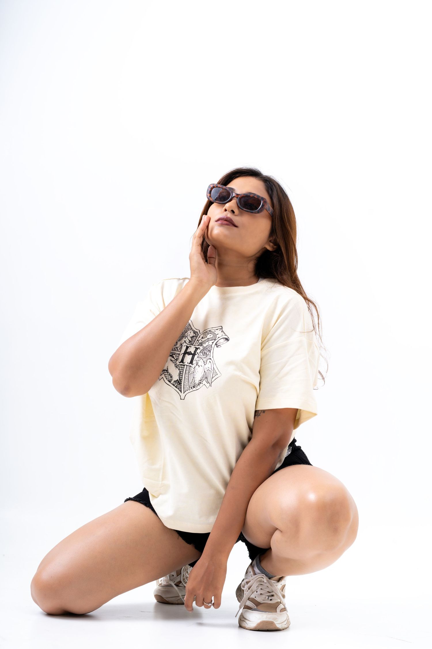 Woman in cream graphic tee, black shorts, and sunglasses posing on knees against white background.