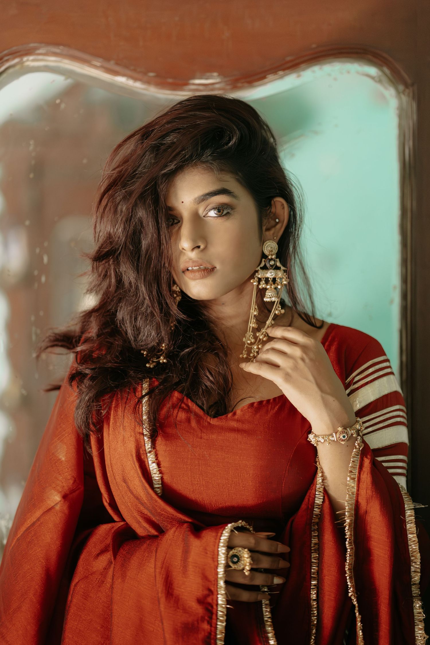 Young woman in red traditional Indian salwar suit with gold chandelier earrings and bracelet, posing elegantly indoors.