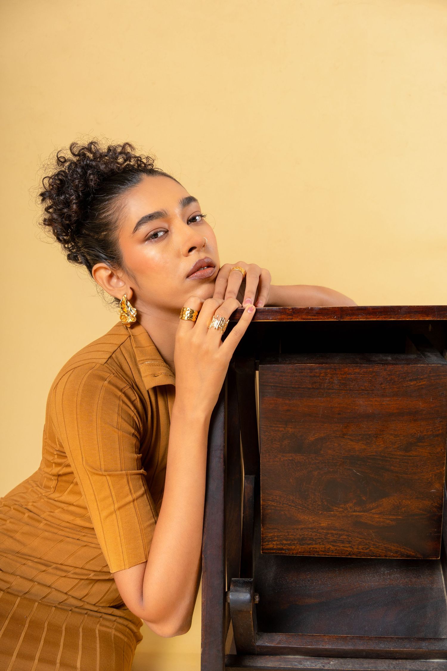 Young woman in mustard dress with gold jewelry leans gracefully against wooden dresser against yellow background.