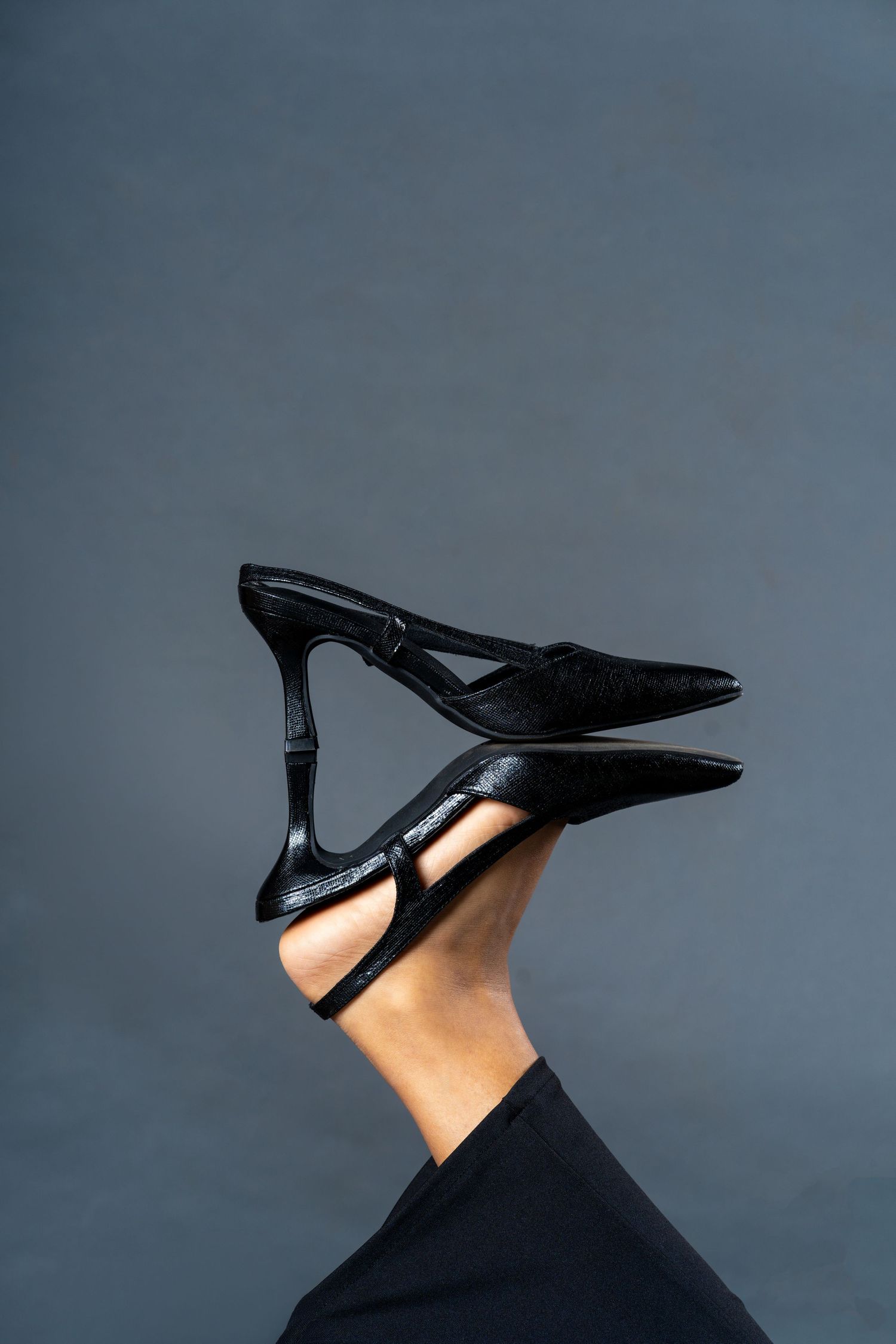 Black pointed-toe stiletto mule heels displayed on raised legs against a gray background.