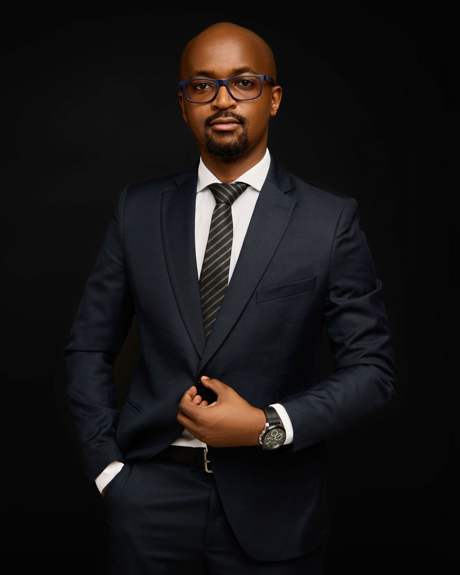 Professional Black man in navy blue suit and glasses posing confidently against dark background.