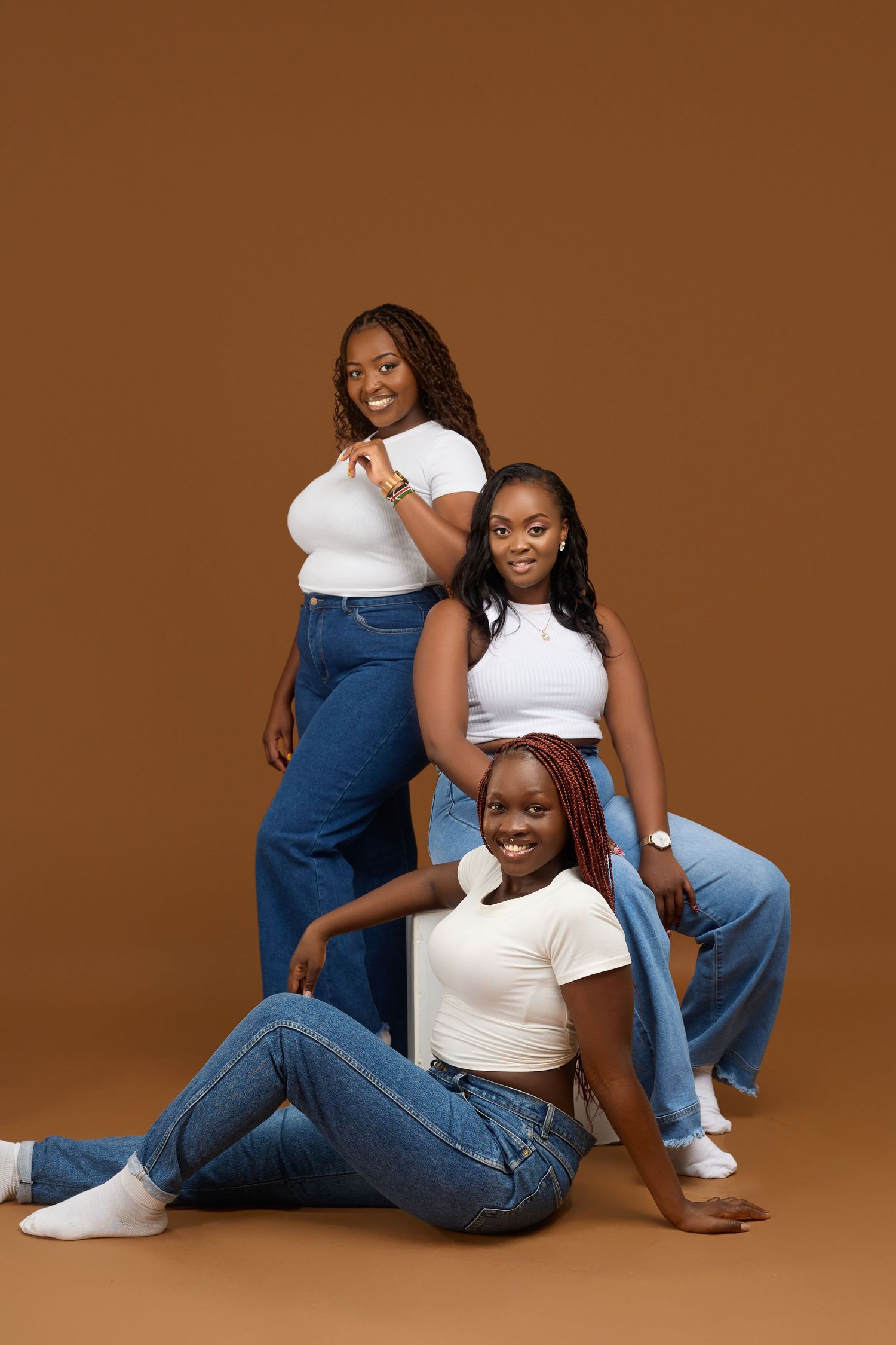 Three young Black women in white tops and blue jeans posing together on brown background.
