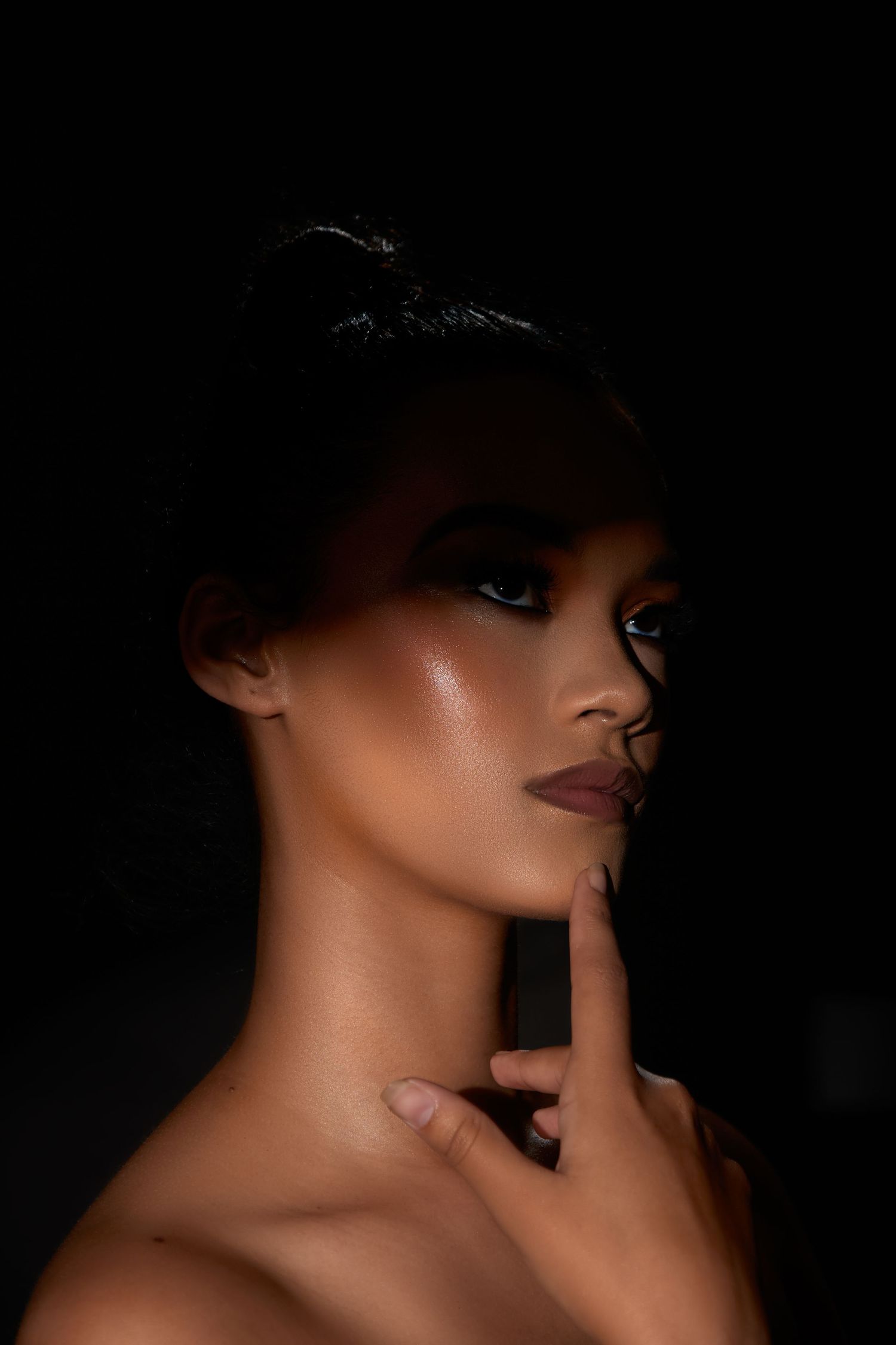A close-up dramatic portrait of a woman with glowing skin and bold makeup posed against a black background.