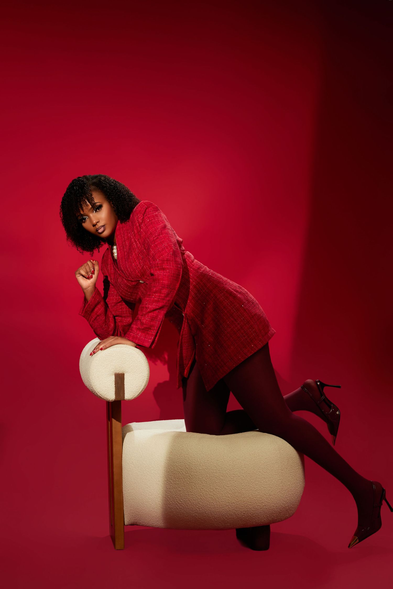 A Black woman in a red tweed jacket and tights leans on a white chair against a bold red background.