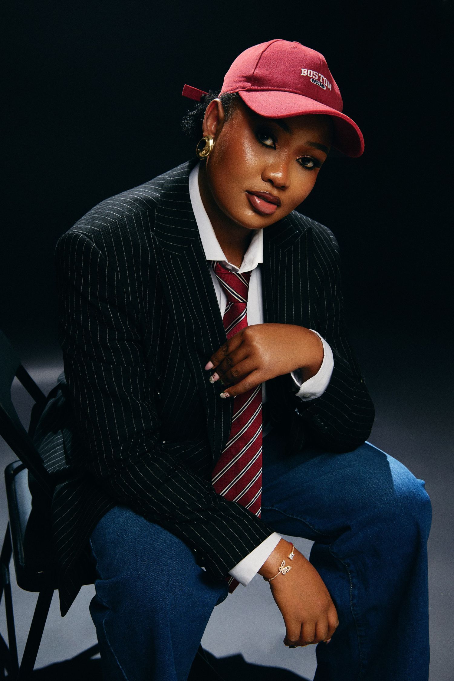 Young woman in pinstripe blazer, red cap, striped tie, and jeans poses on chair against dark background.