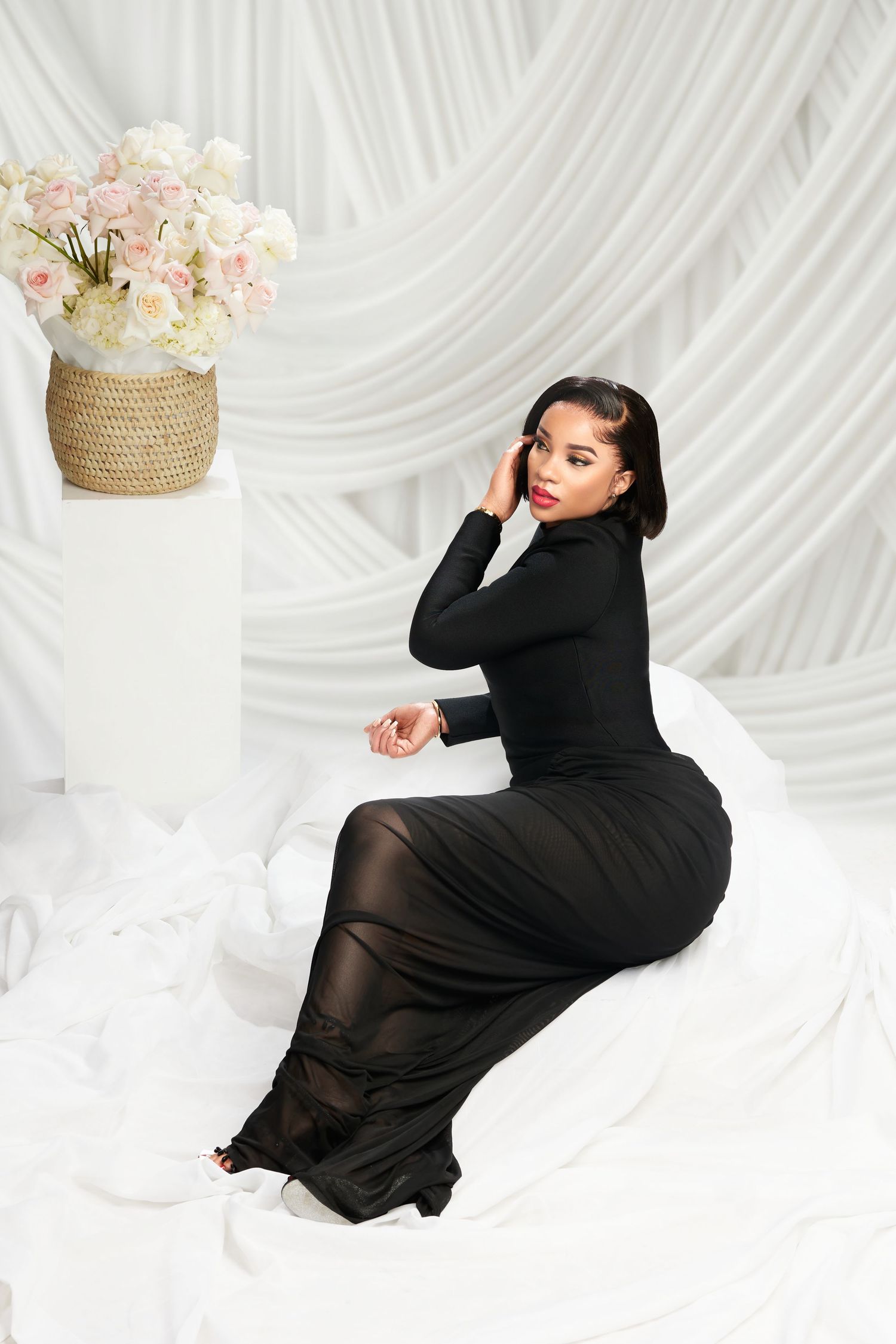 Woman in black turtleneck maxi dress sits gracefully on white fabric beside pink roses in wicker vase.