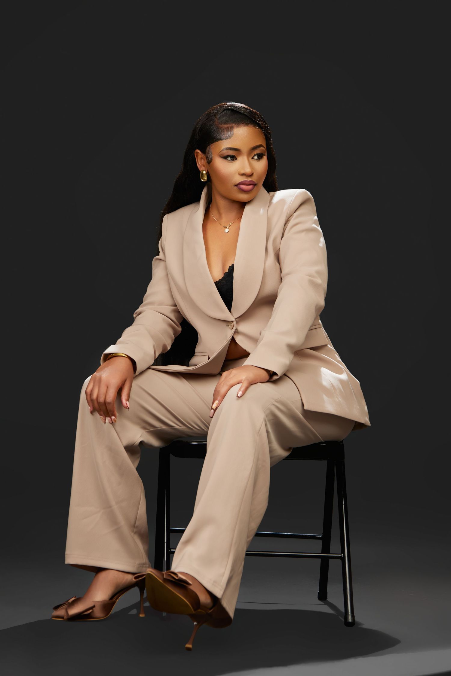 A Black woman sits confidently on a stool wearing a beige blazer suit with a black camisole underneath.