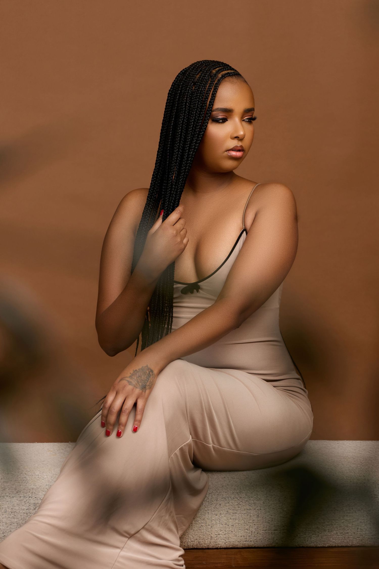 A woman with long box braids sits elegantly in a beige form-fitting outfit against a warm brown background.