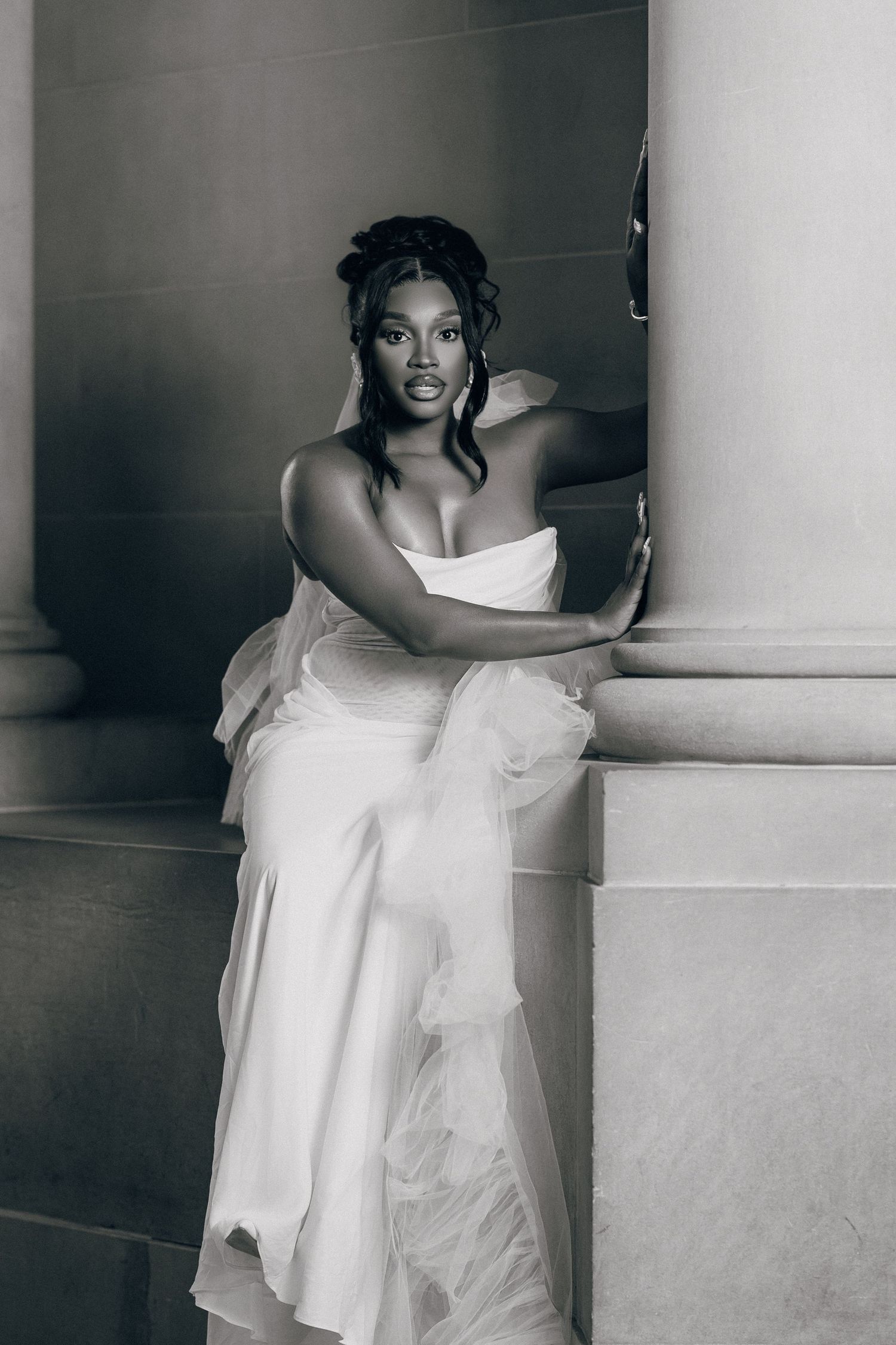 Black and white portrait of a bride in a strapless gown with tulle detail, leaning confidently against a stone column.
