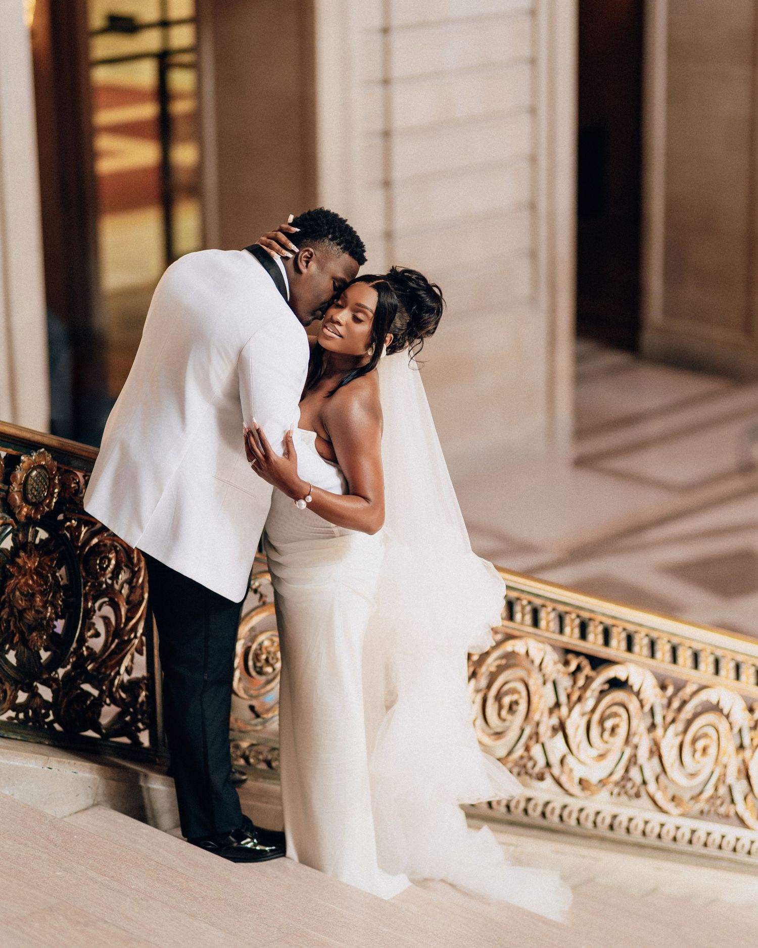 Groom in a white dinner jacket embraces his bride in a strapless gown and veil beside an ornate gold railing.