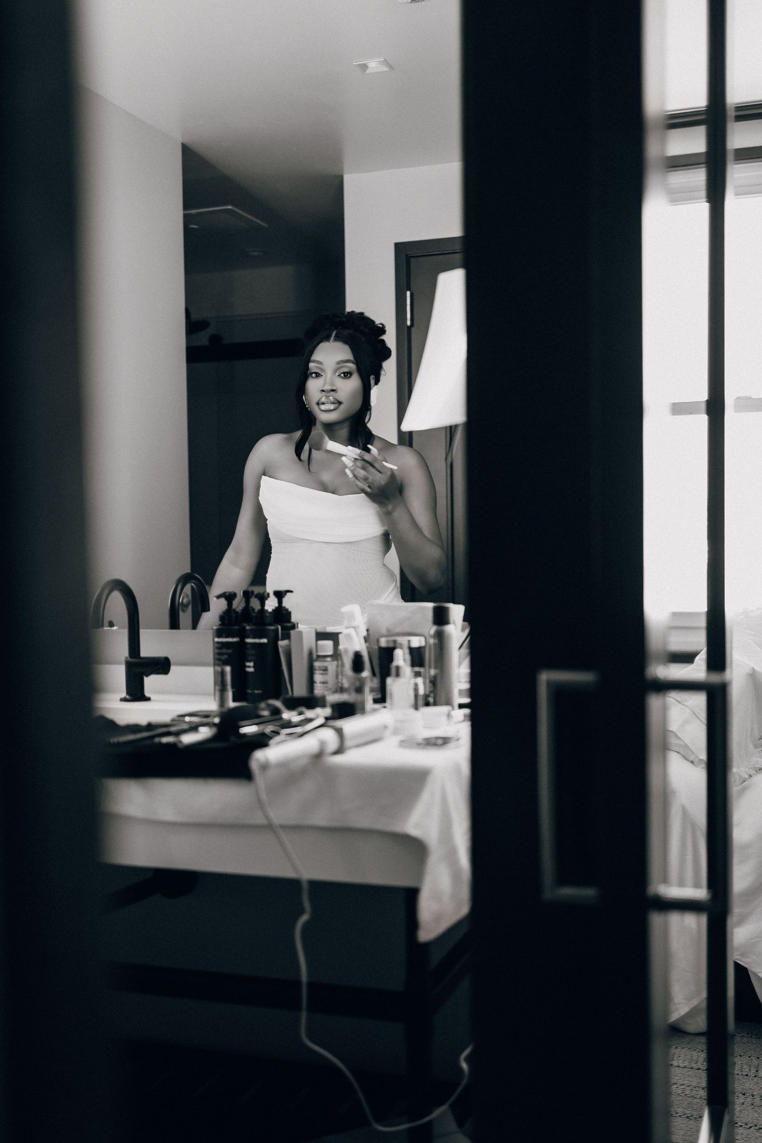 Black and white photo of a bride in a towel-style wrap getting ready, reflected in a mirror beside a vanity of beauty products.