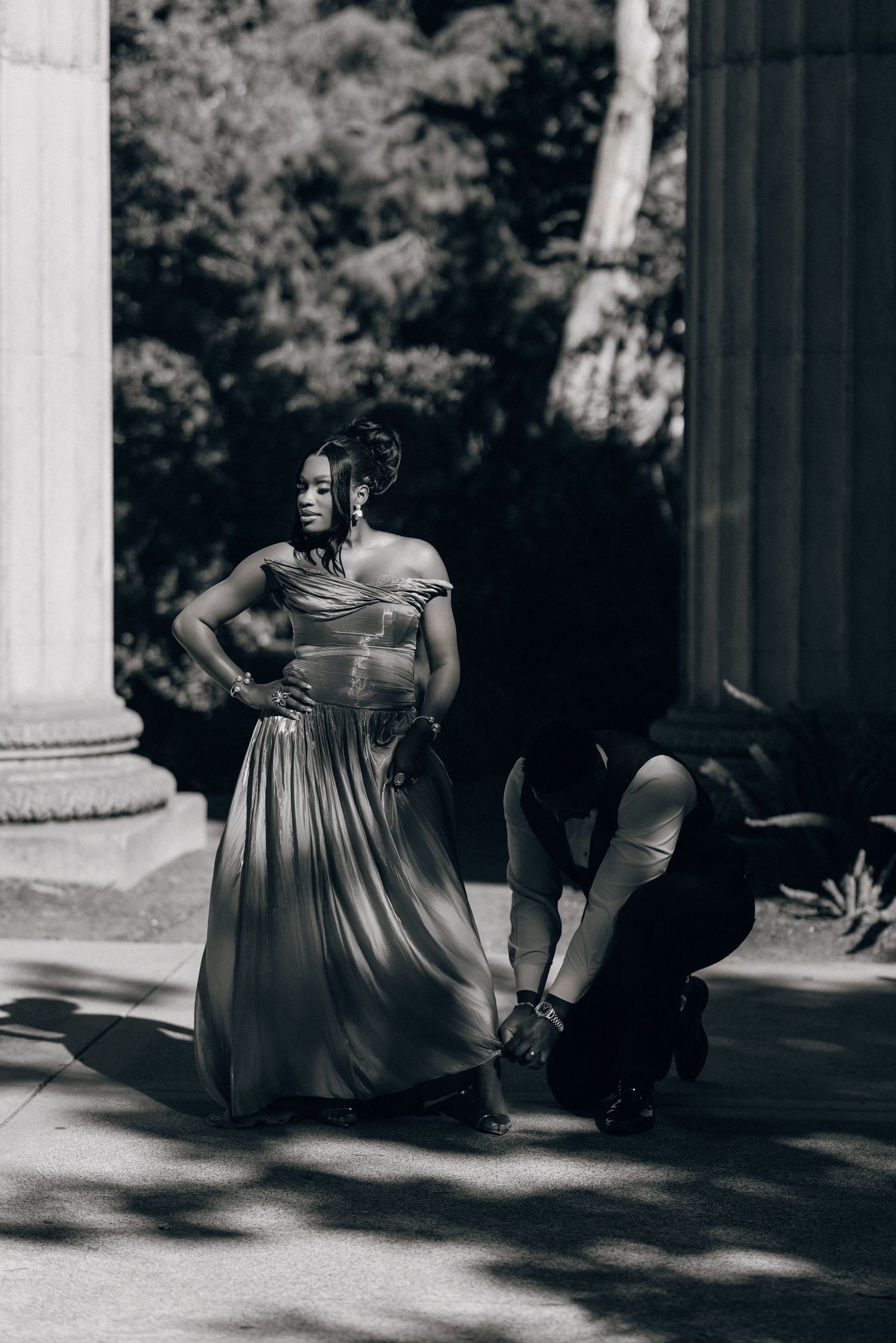 Black and white photo of a woman in a flowing satin gown posing as her partner kneels to adjust his shoe.