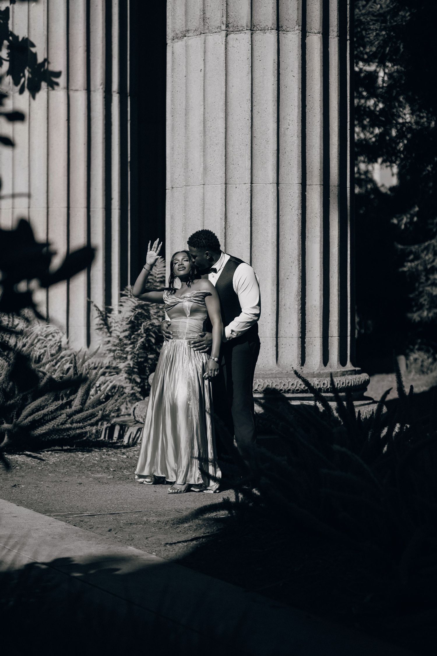 Black and white image of a man kissing his partner's cheek as she raises her hand by a stone column.