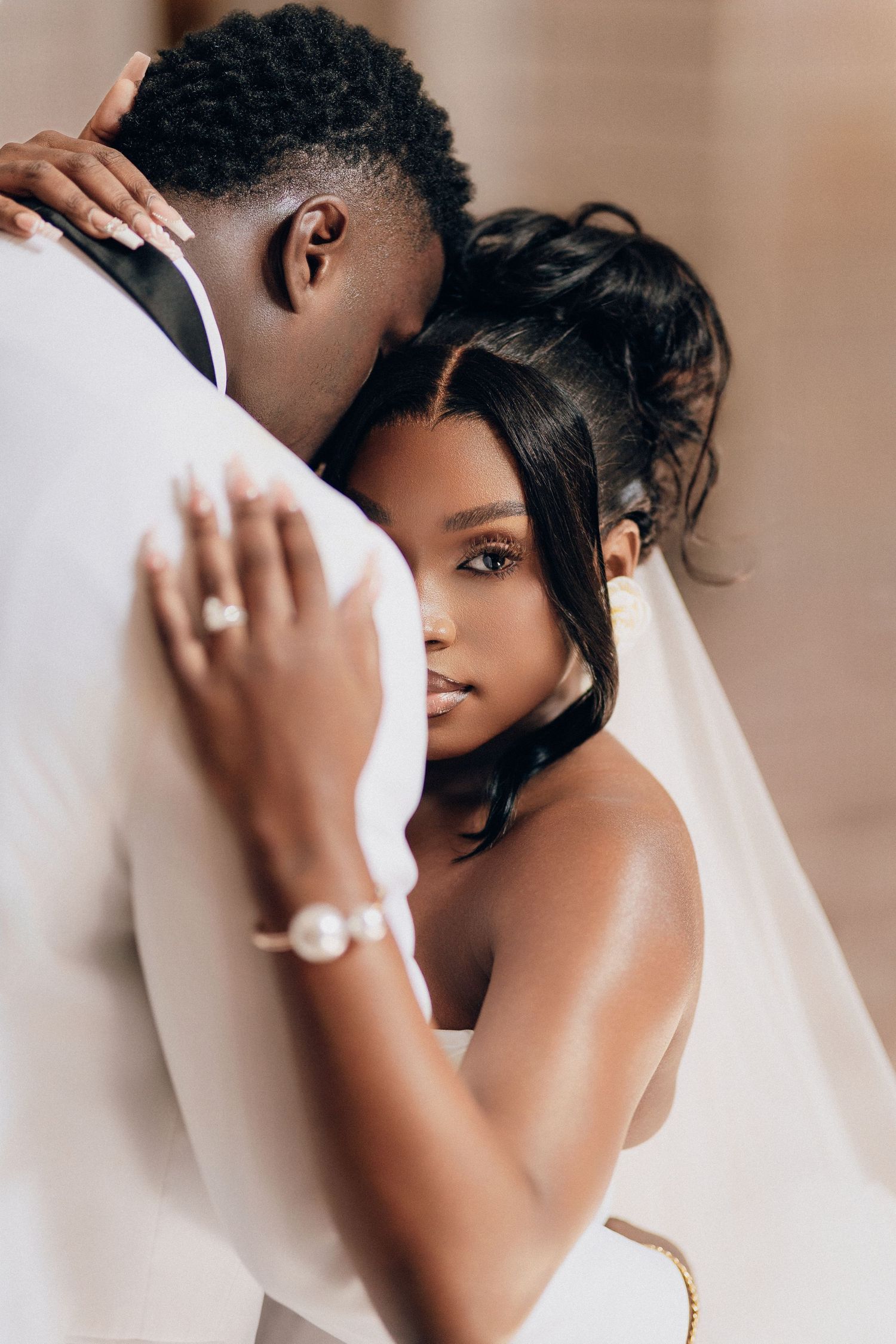 Elegant bride in a veil and white gown rests against her groom in a tender embrace, her ring and bracelet visible on his shoulder.