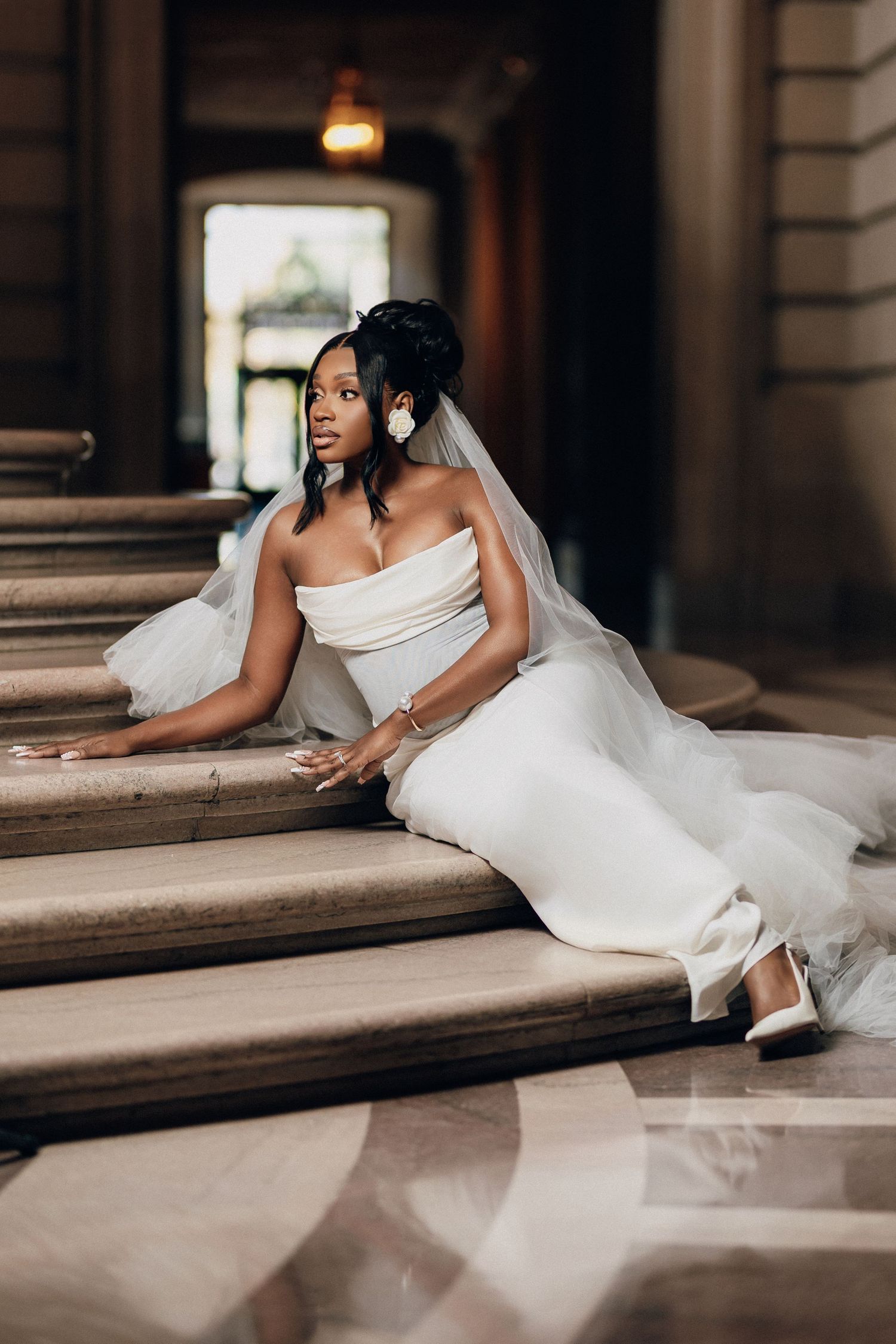 Bride in a strapless white gown and veil sits gracefully on marble steps in a historic building, gazing into the distance.