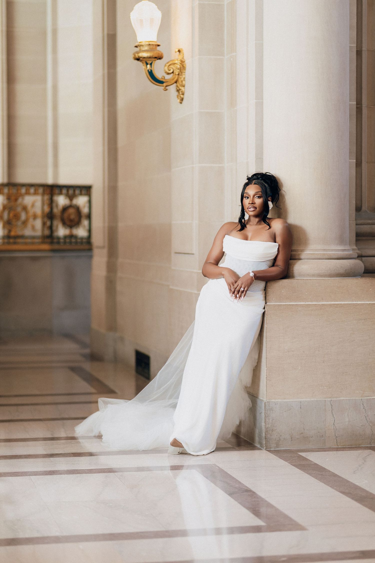 Bride in a strapless white gown with a long veil leans against a column in an elegant marble hallway.