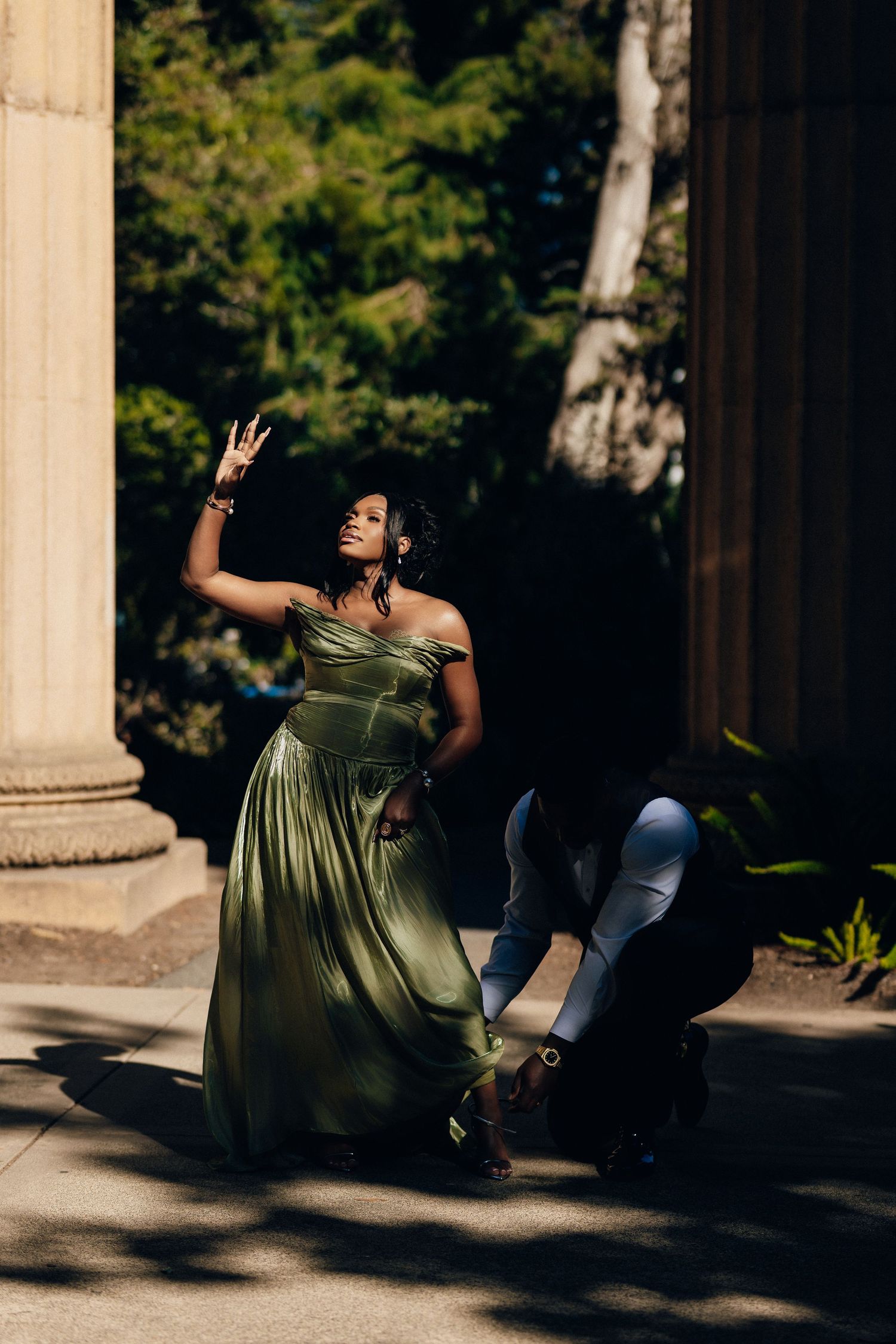 Woman in an olive green satin gown raises her hand toward the sky as her partner kneels nearby among tall columns.