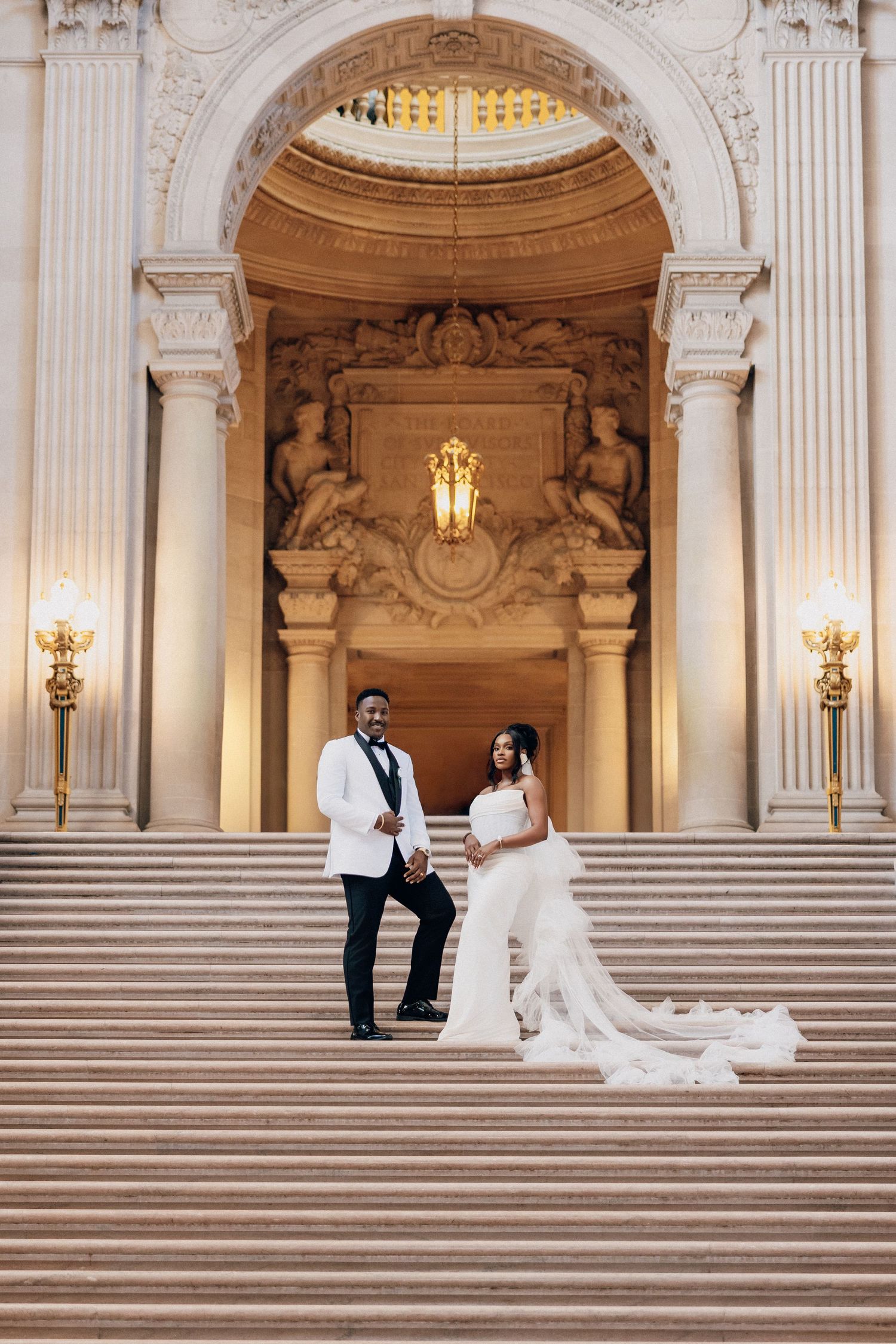 Bride and groom pose on grand marble steps beneath an ornate arch, she in a white gown with a long veil and he in a white tuxedo jacket.