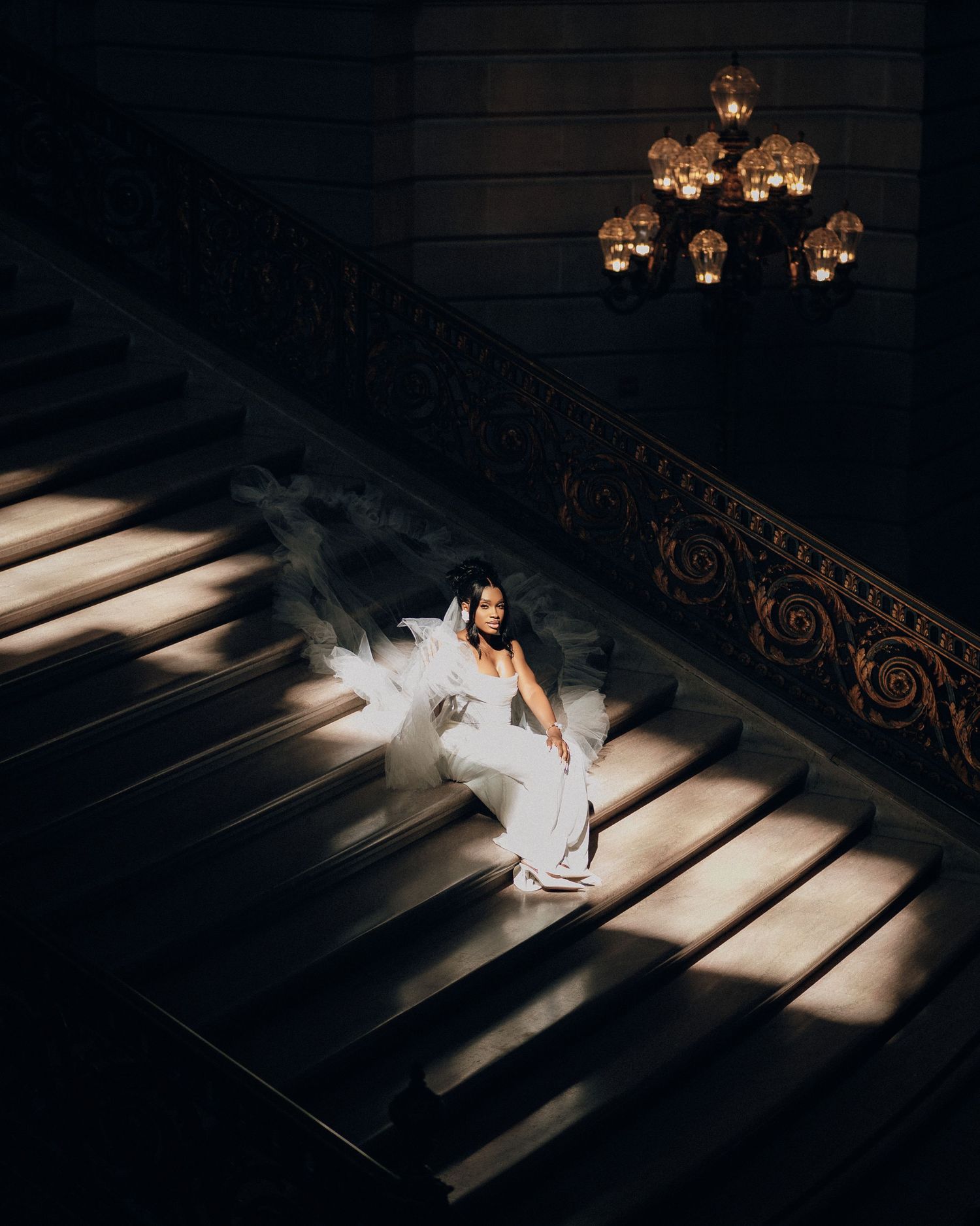Bride in a flowing white gown and veil sits on a grand staircase lit by dramatic sunlight beneath an ornate chandelier.