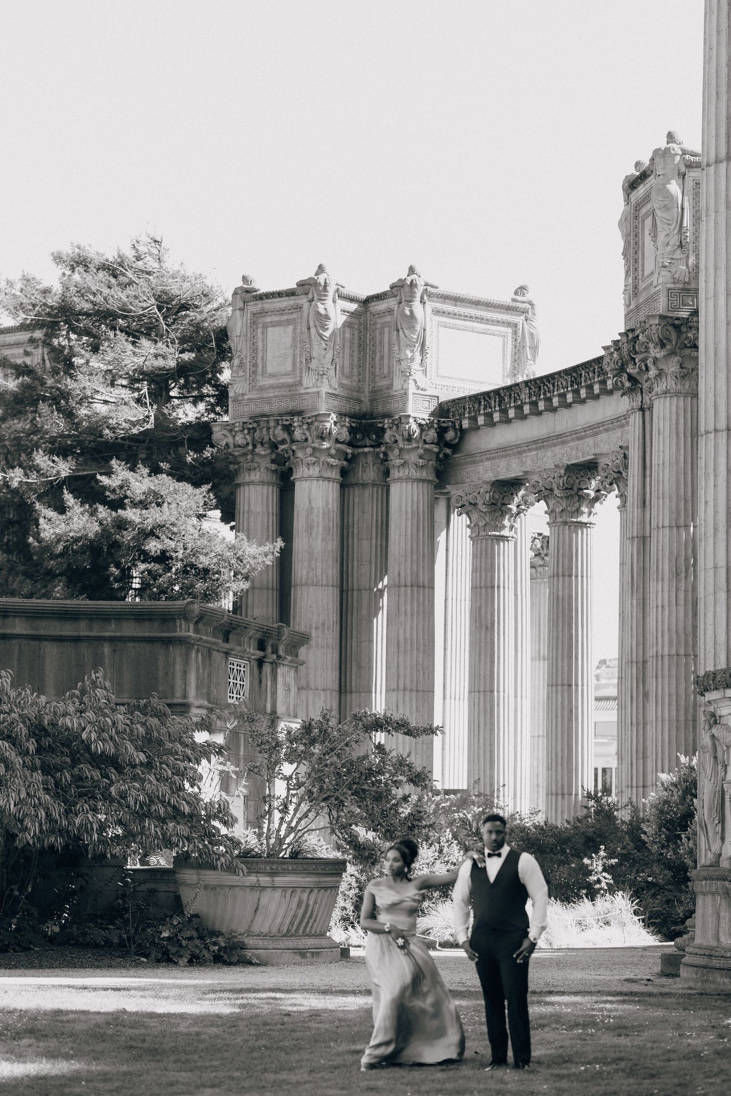 Black and white photo of a couple walking hand in hand before a classical colonnade surrounded by trees.