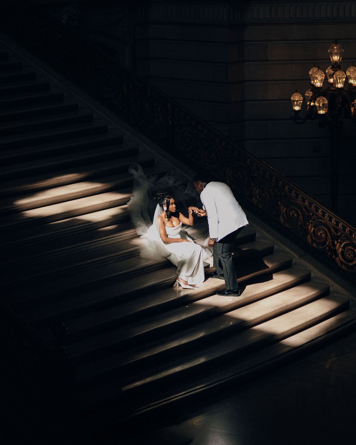 Bride in a white gown and veil sits on a grand staircase as her groom bends toward her in dramatic light.