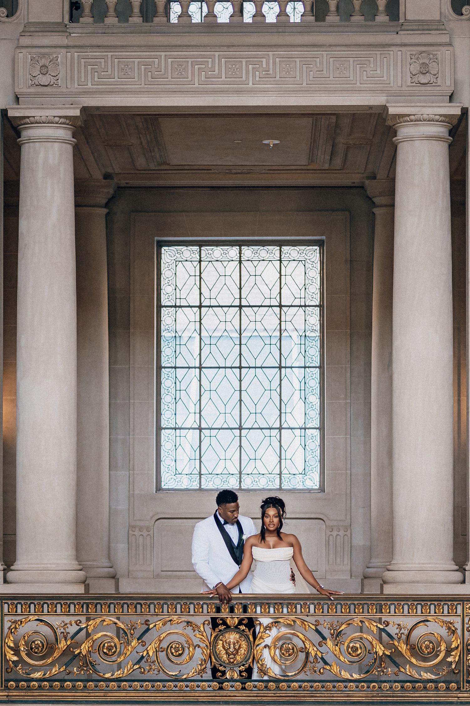 Couple in wedding attire stand on a gilded balcony beneath a large lattice window framed by stone columns.