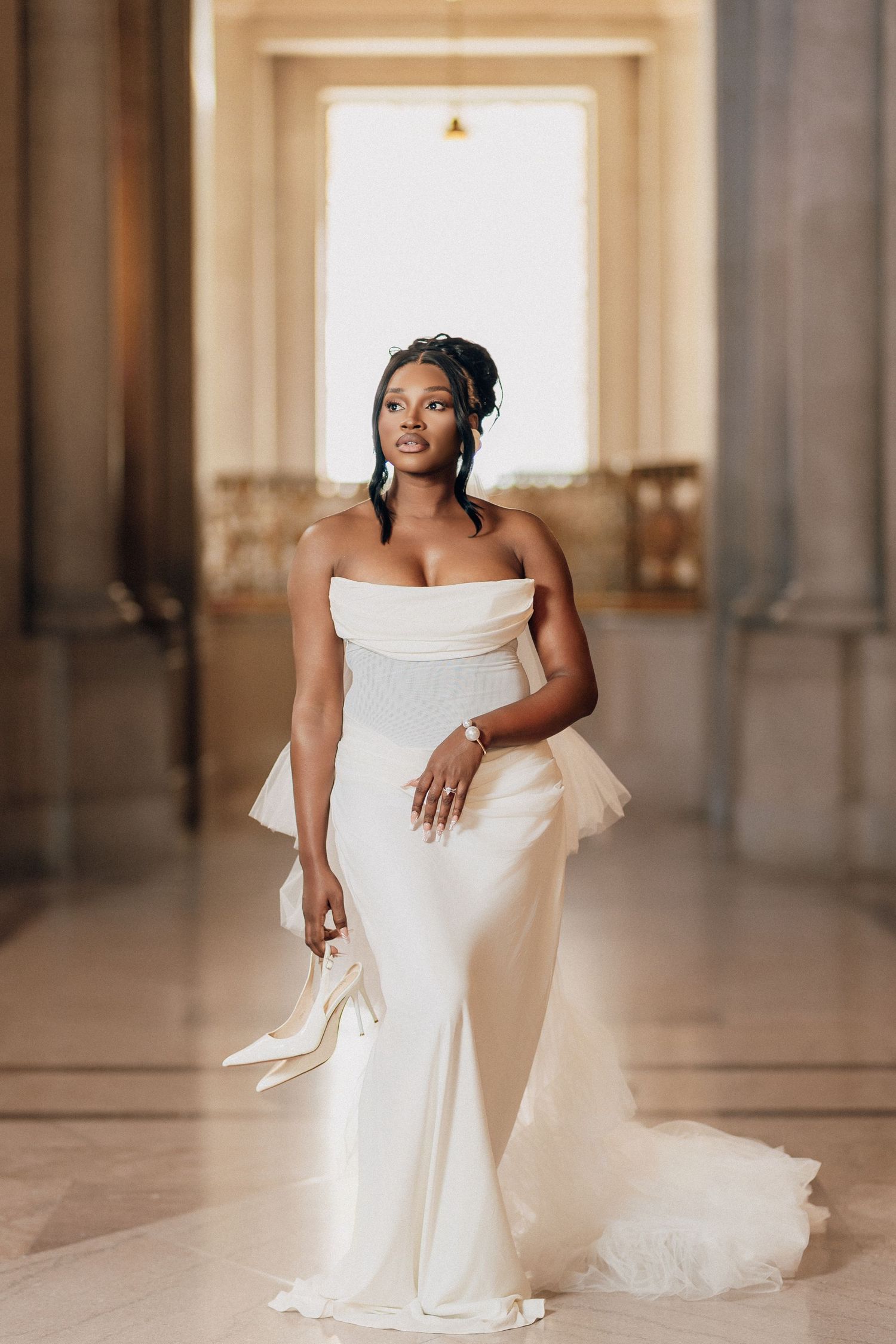 Bride in a fitted strapless gown holds her heels while standing in a sunlit marble hall.