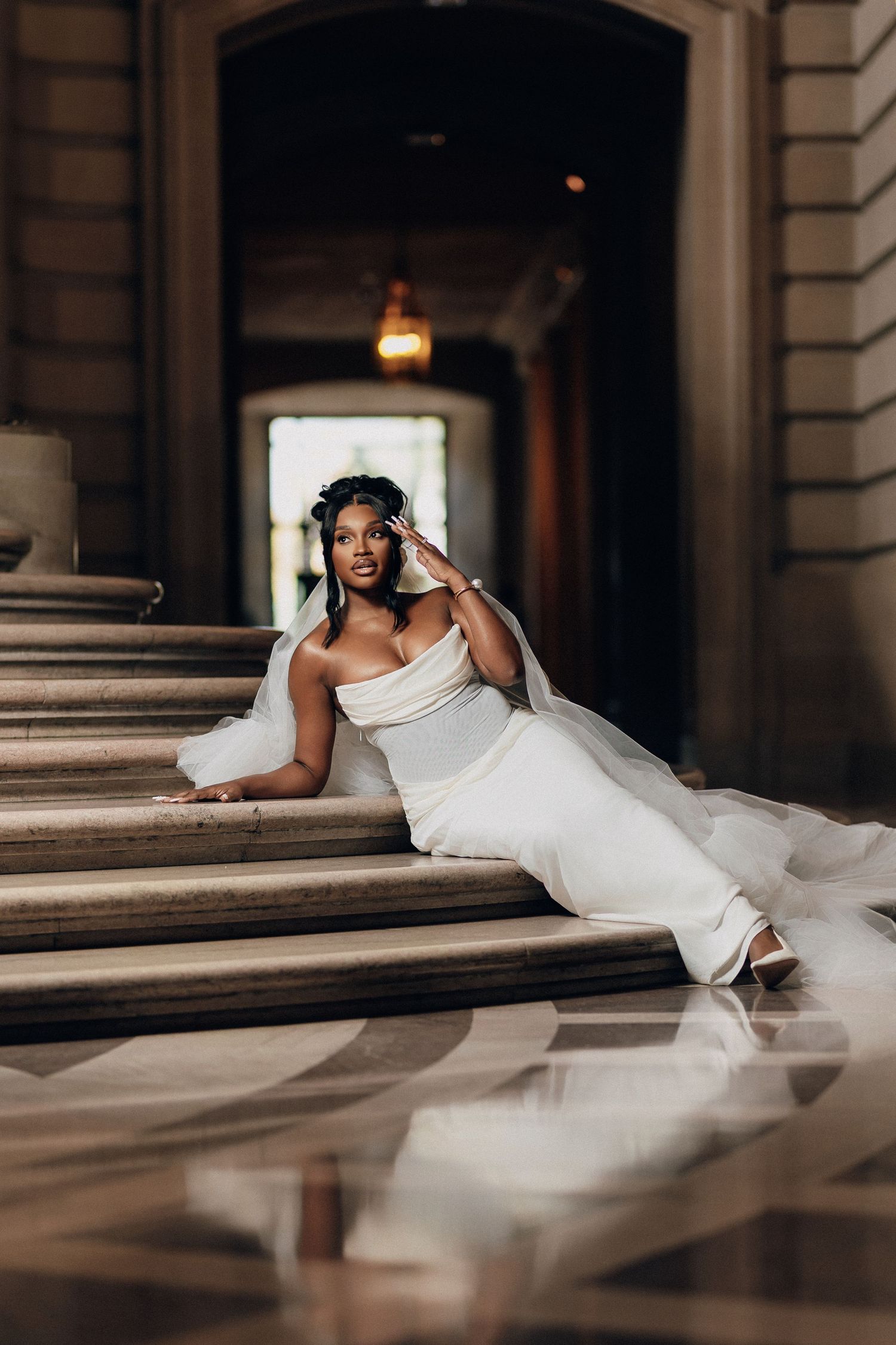 Bride in a white gown and sheer veil reclines on grand stairs, touching her face, with warm lights glowing in the background.