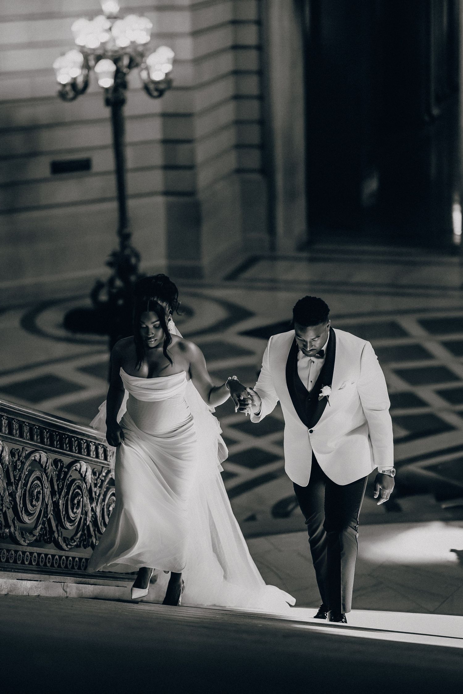 Bride in a strapless white gown and groom in a white tuxedo jacket hold hands walking down ornate marble stairs in black and white.