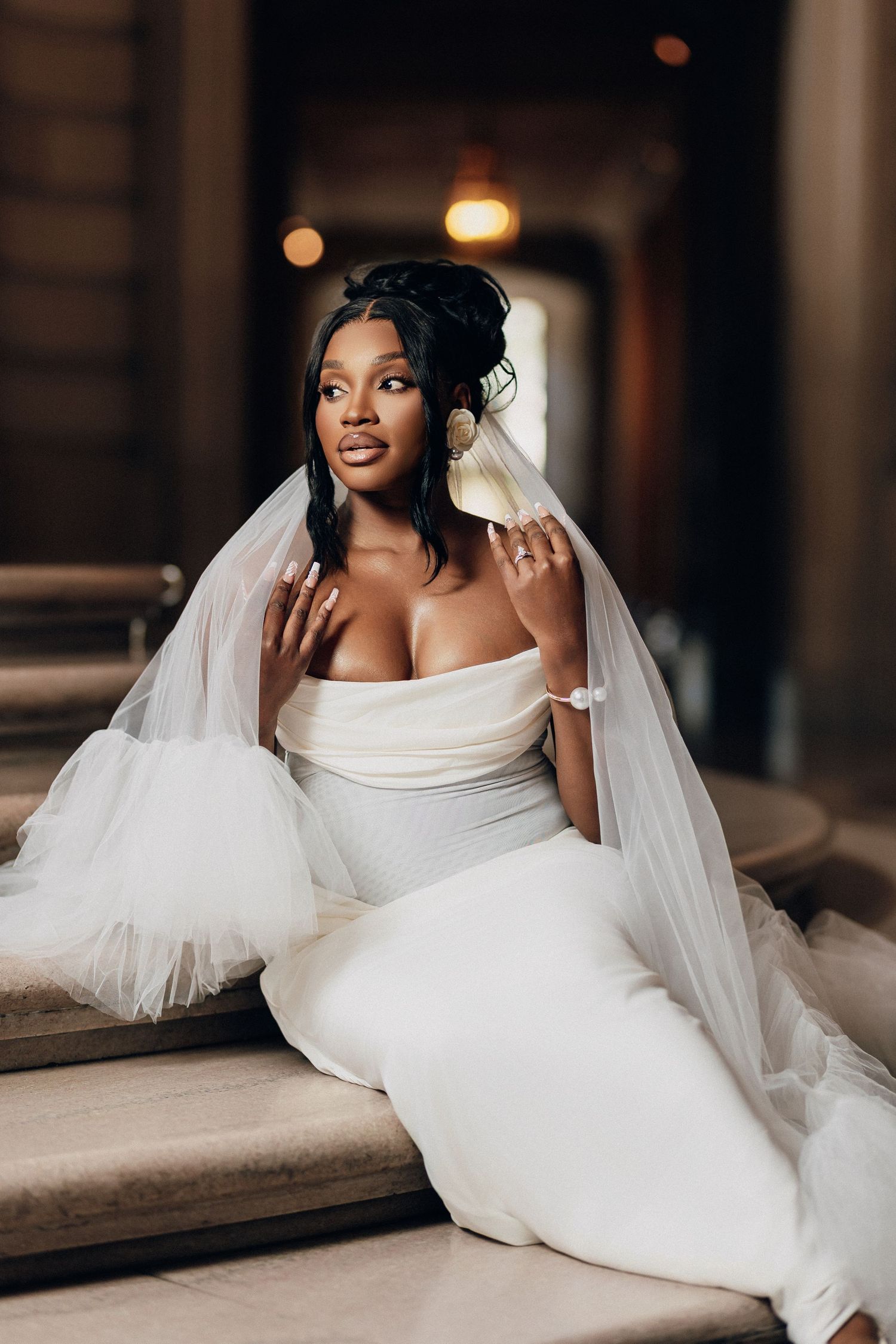 Smiling bride with an elegant updo and a tulle veil sits on marble steps in a strapless white gown, framed by soft golden lights.