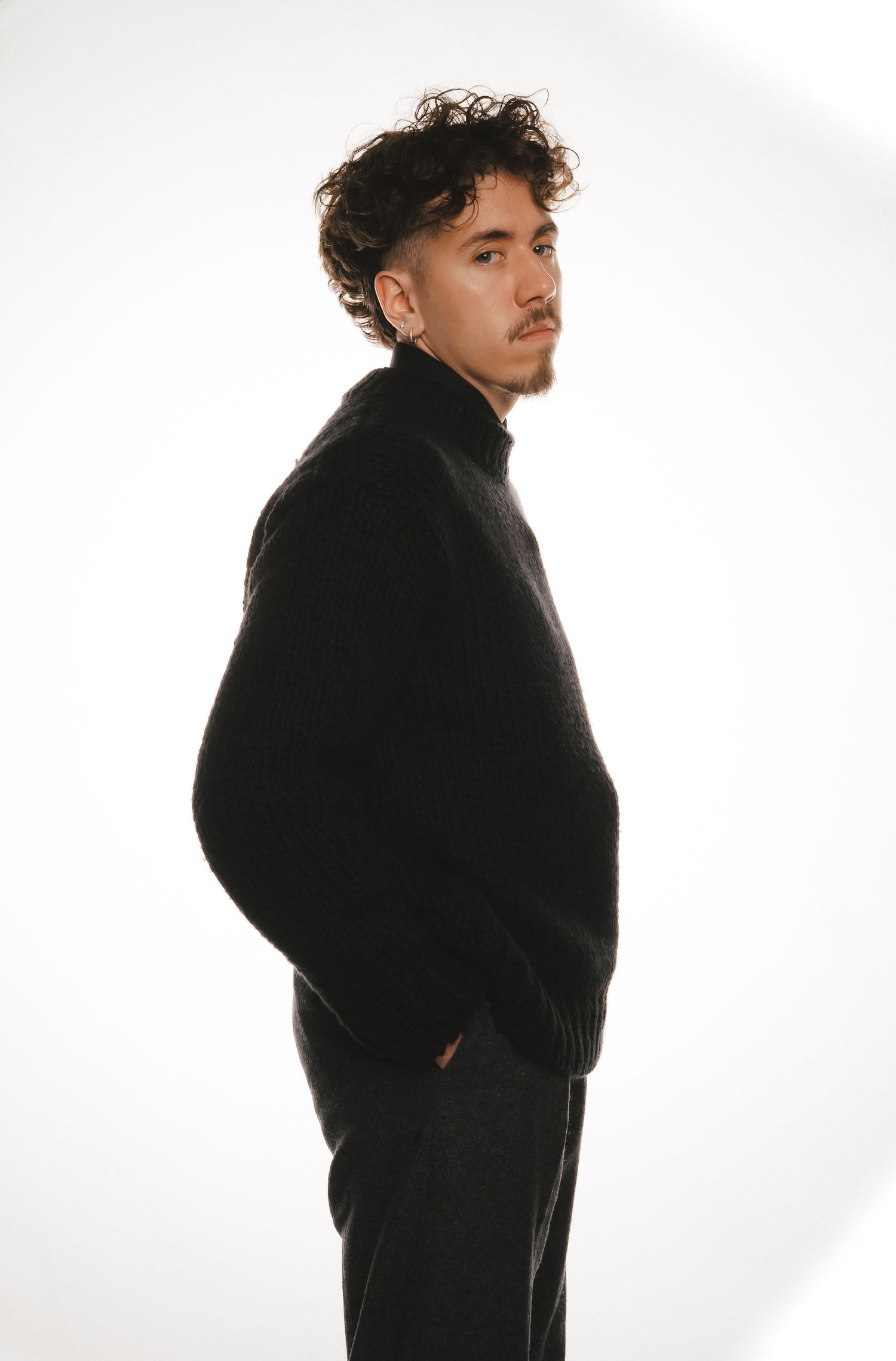 Side profile of young man with curly hair in black turtleneck sweater posing against white background.