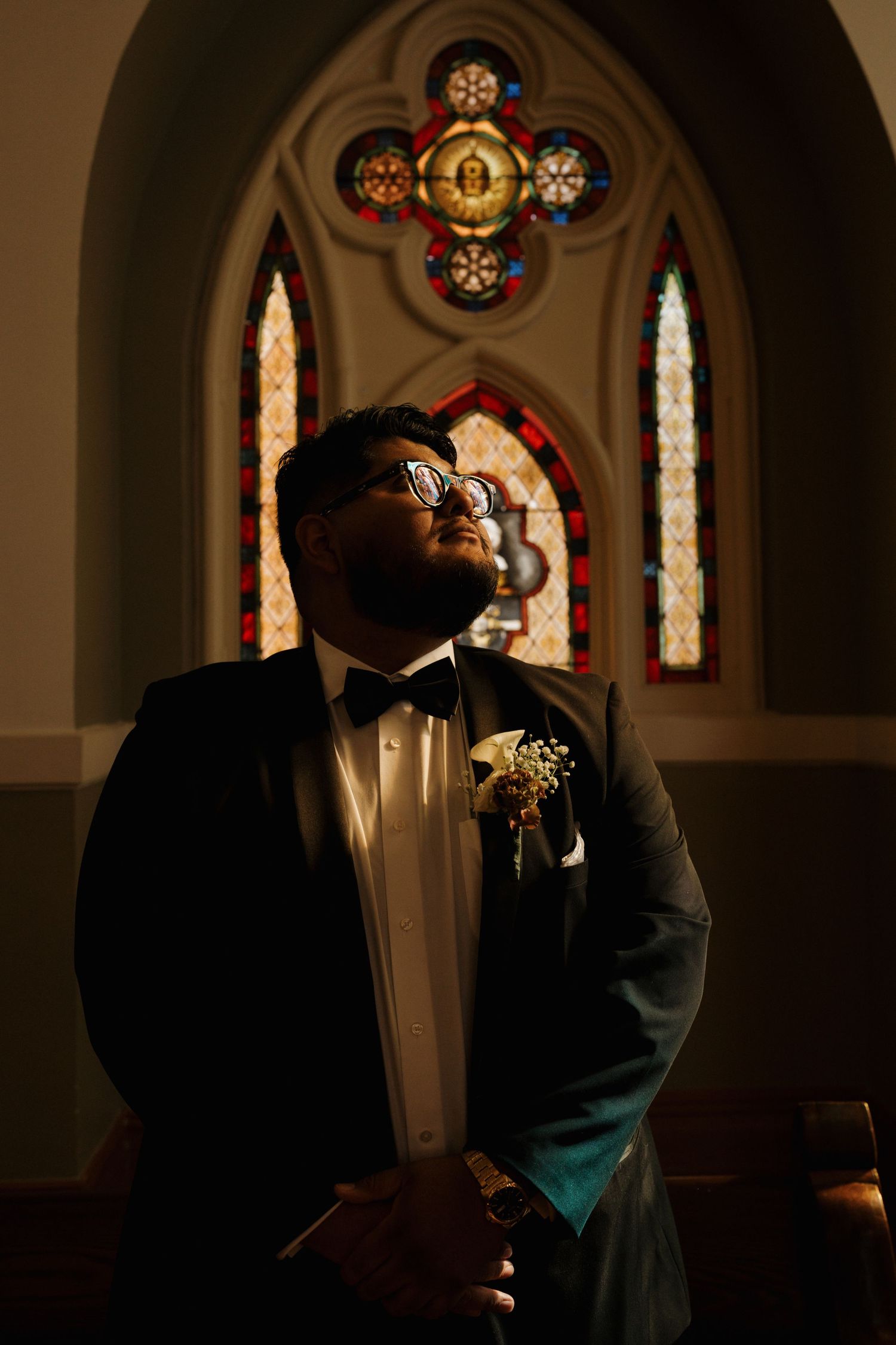 Groom in black tuxedo with boutonniere standing beneath colorful stained glass church windows.