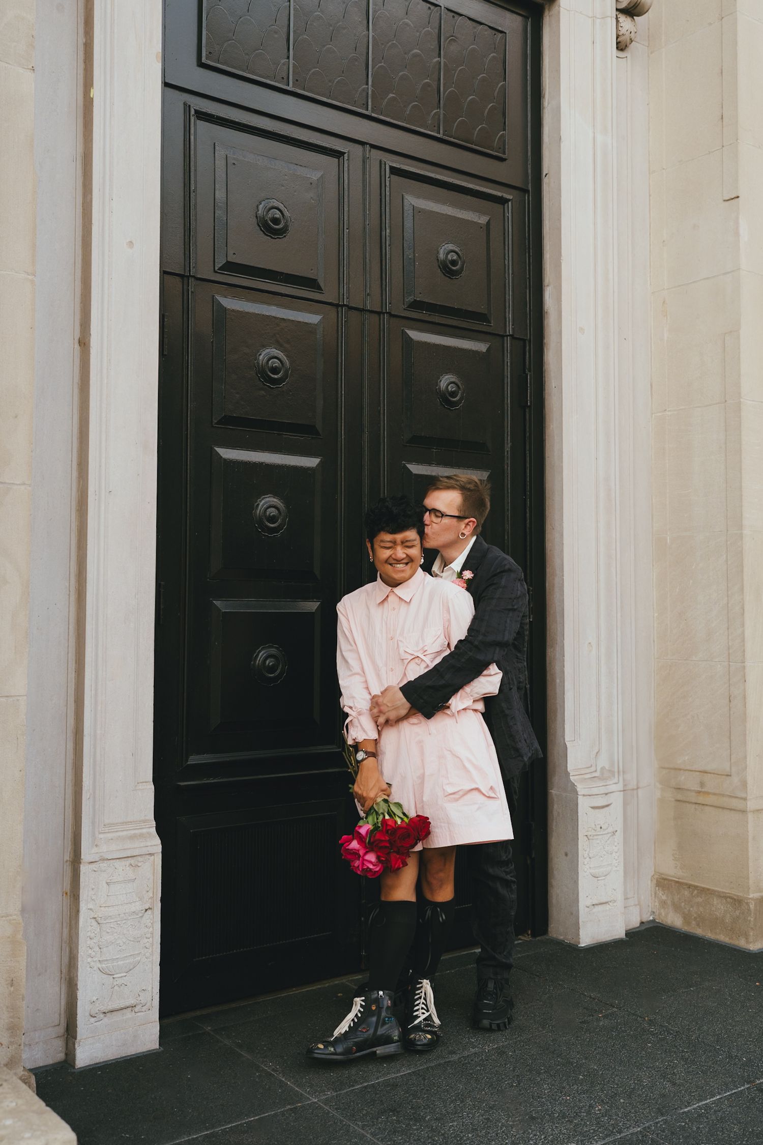 Man in dark suit embracing woman in pink dress holding red roses, standing before grand black double doors.