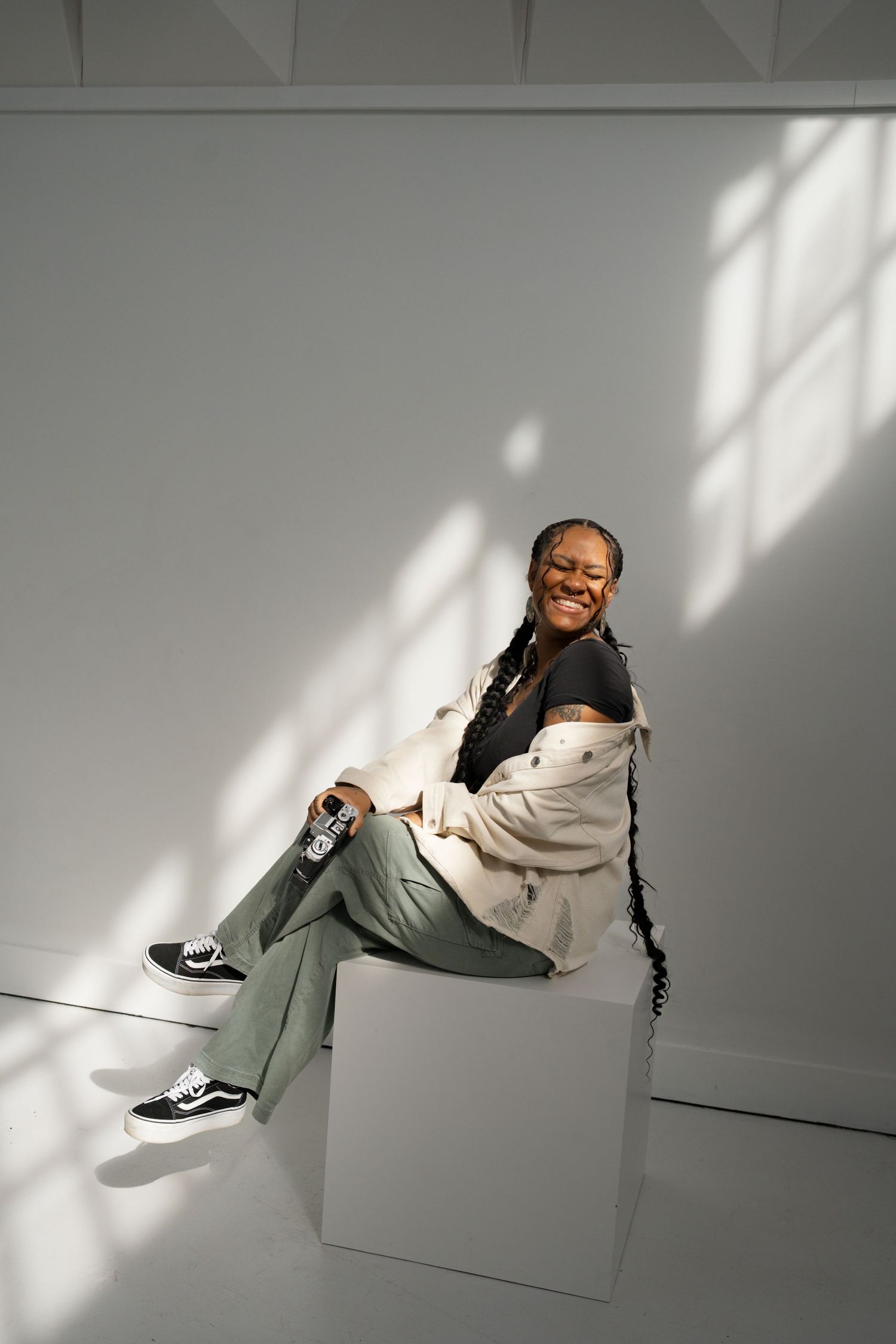 A Black woman with braids sits on a white cube, smiling, wearing a white jacket, black top, sage green pants, and Vans sneakers.