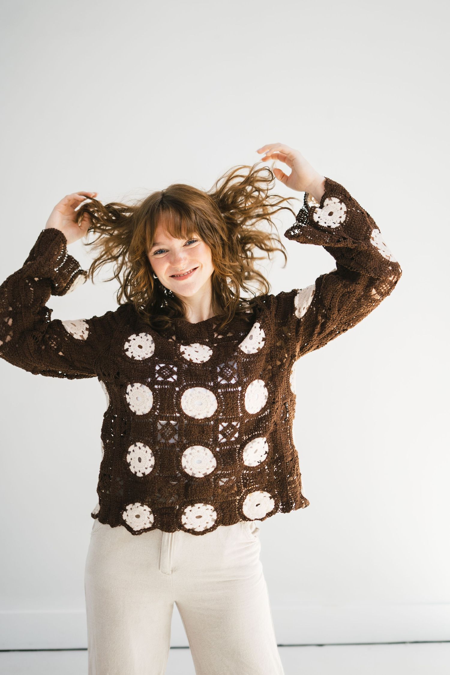 A smiling woman with tousled hair wears a brown crochet granny square sweater with white floral motifs.