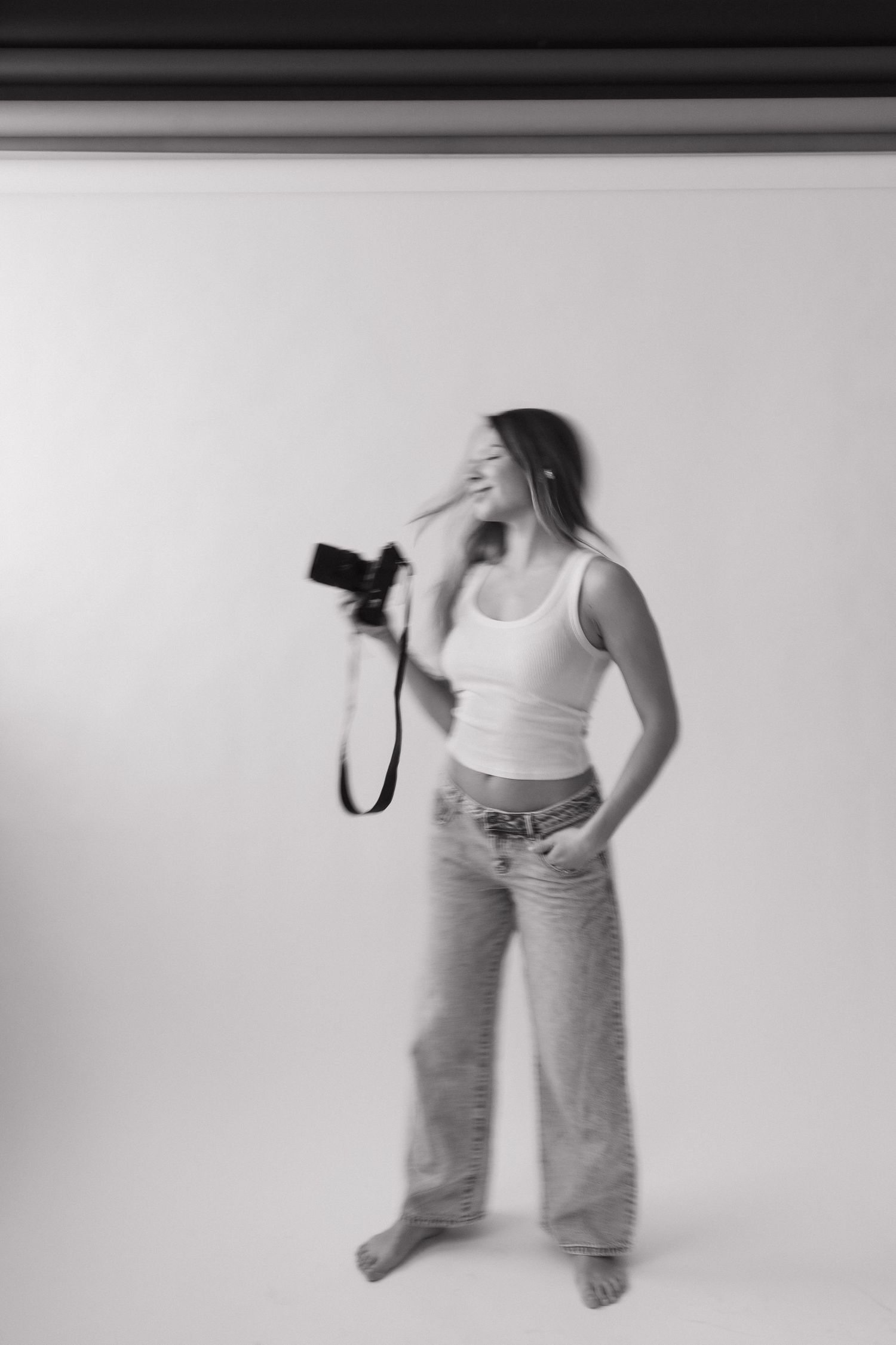 Black and white motion blur photo of smiling woman in tank top and jeans holding camera in studio.
