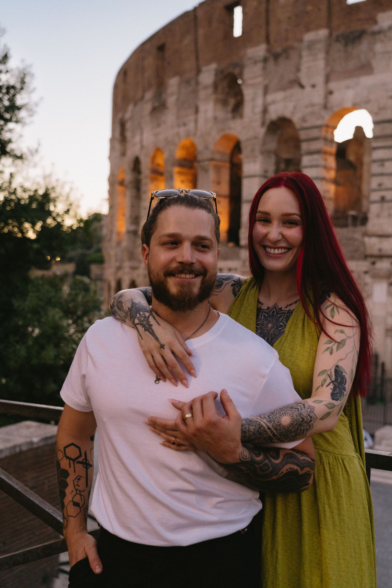 Tattooed couple embracing and smiling in front of the illuminated Colosseum in Rome at dusk.