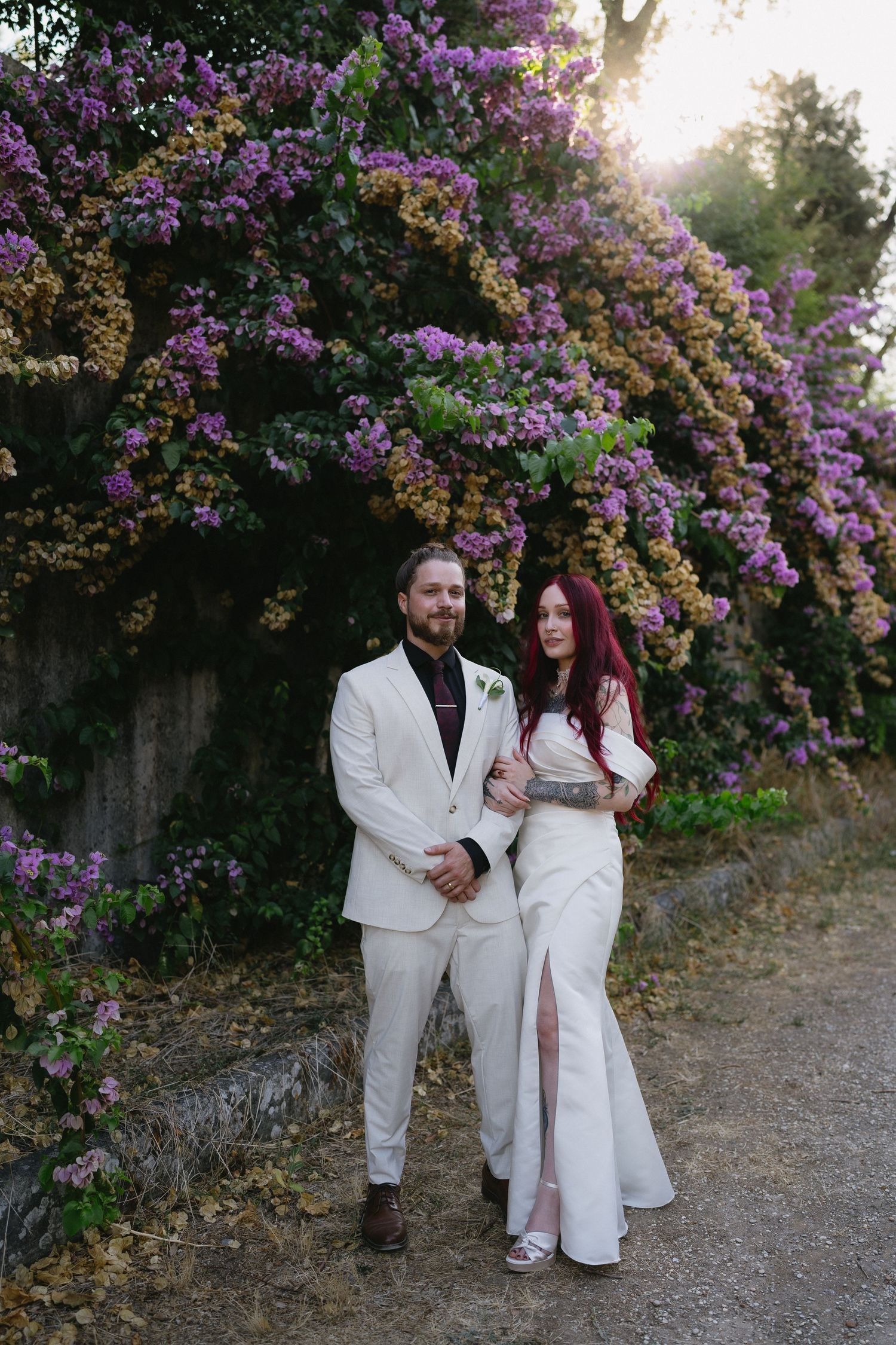 Wedding couple posing before vibrant purple bougainvillea at golden hour in romantic garden setting.