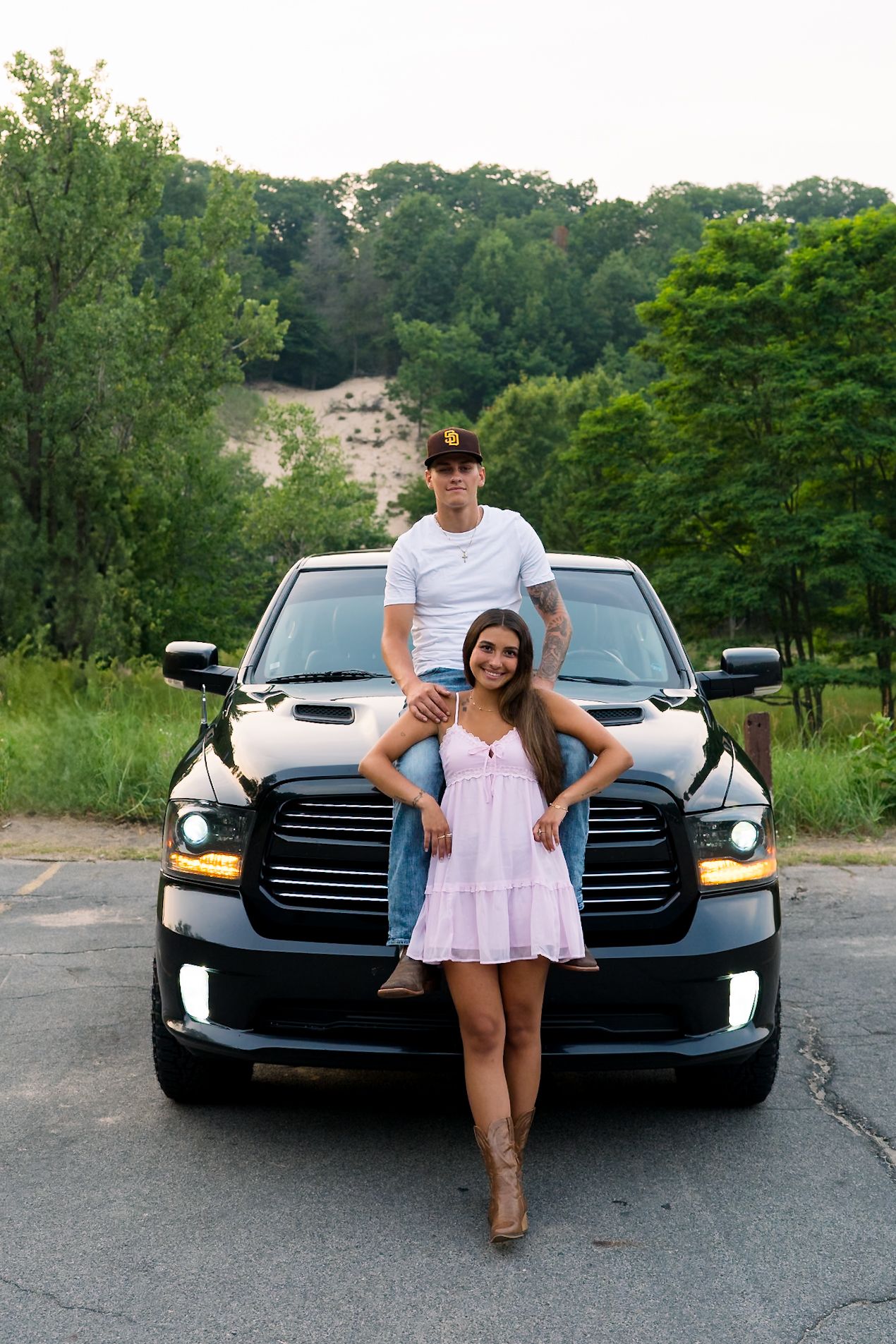 Young couple posing with black Dodge Ram truck, man standing on bumper embracing woman in pink dress and boots.