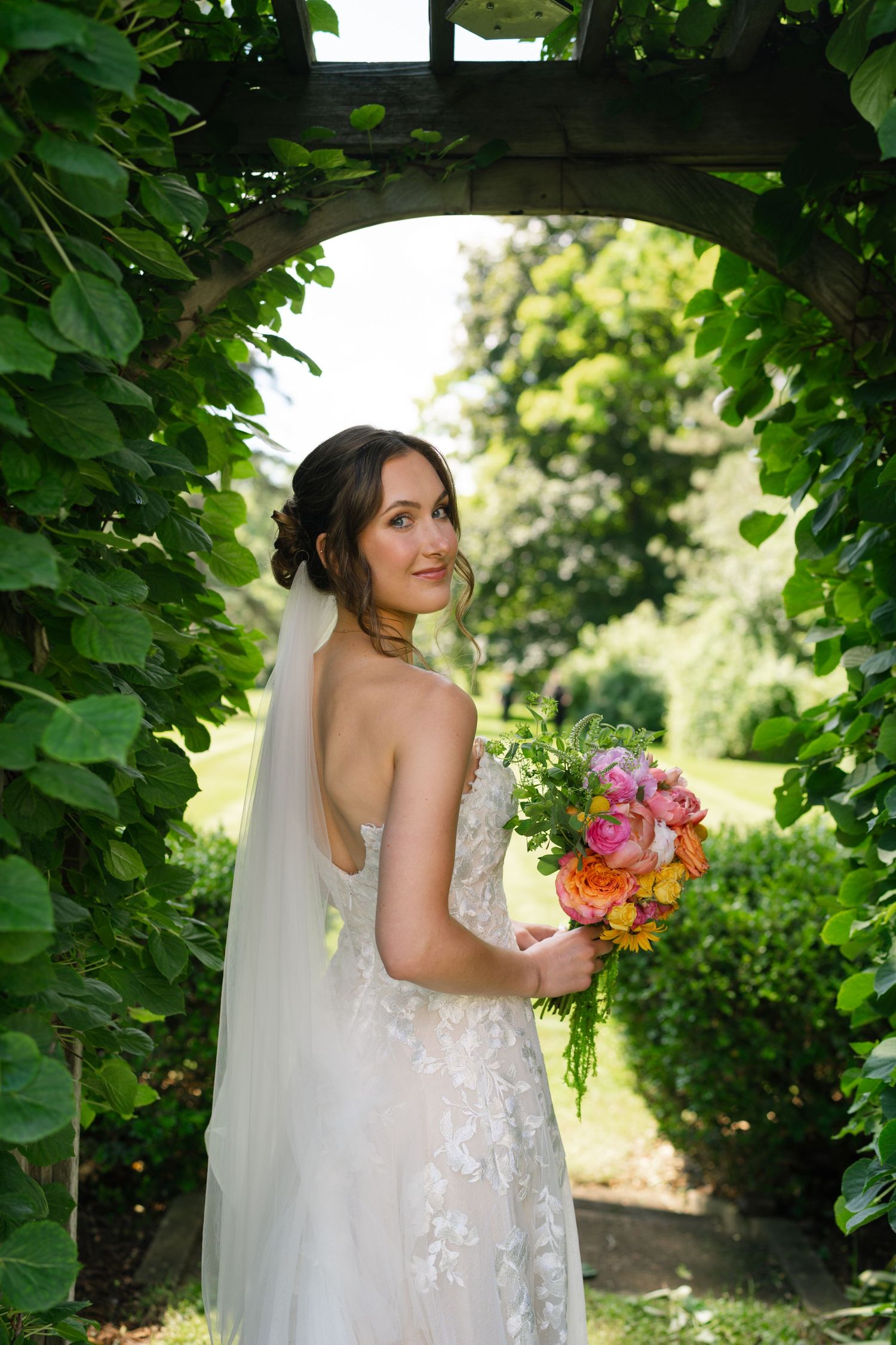 Bride in a lace wedding gown holding a colorful bouquet standing under a green leafy garden arch.