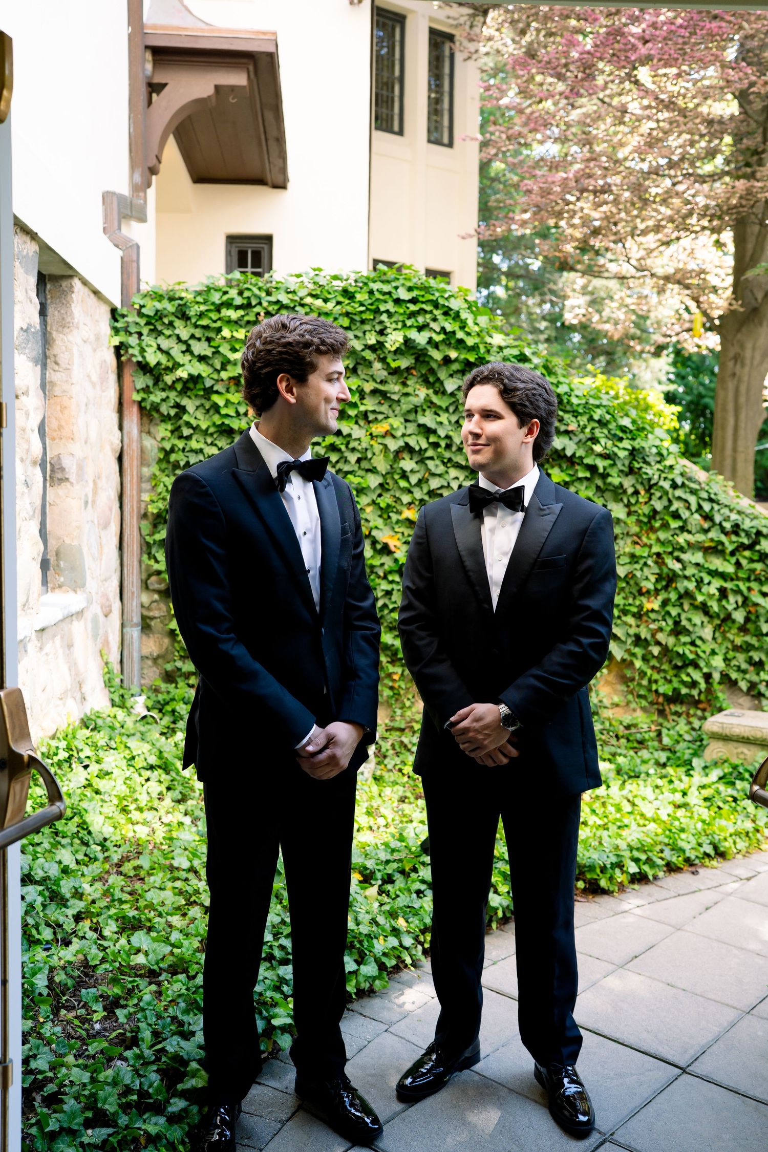 Two grooms in black tuxedos with bow ties share a smile outdoors against an ivy-covered wall.