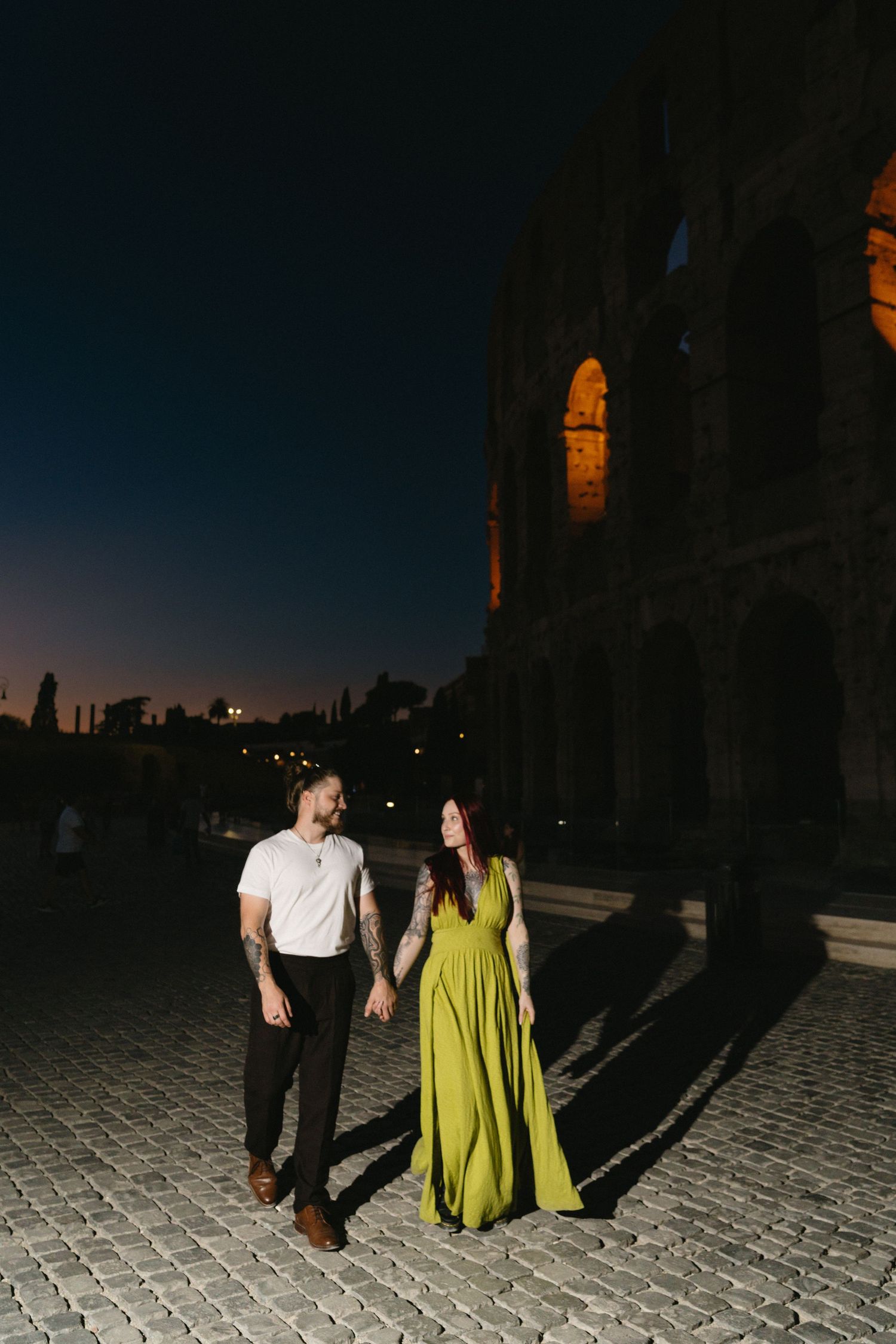 Couple holding hands at night near the illuminated Colosseum in Rome, woman in green gown, man in white shirt.
