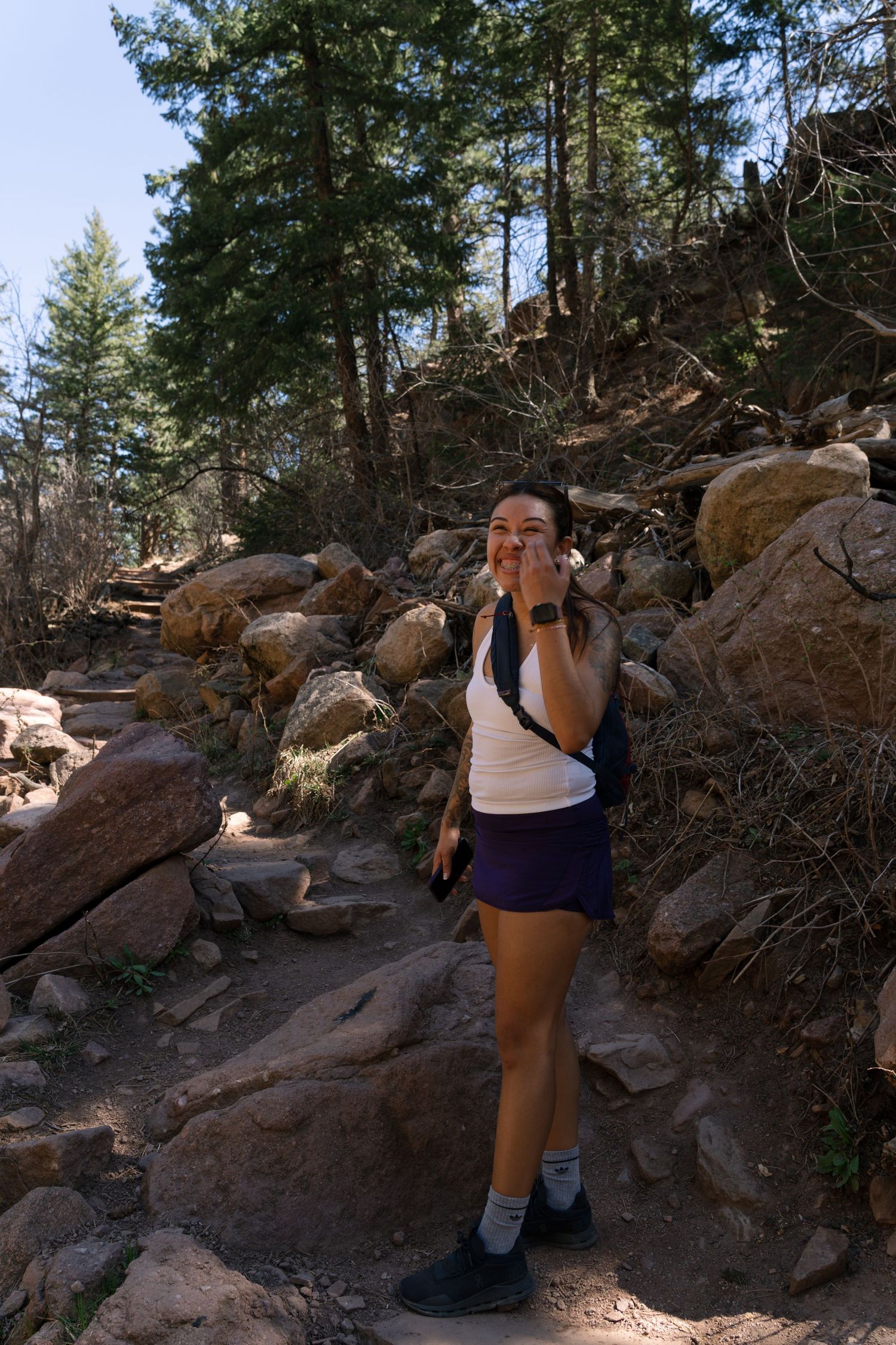 Smiling woman hiker poses on a rocky mountain trail surrounded by pine trees and boulders.