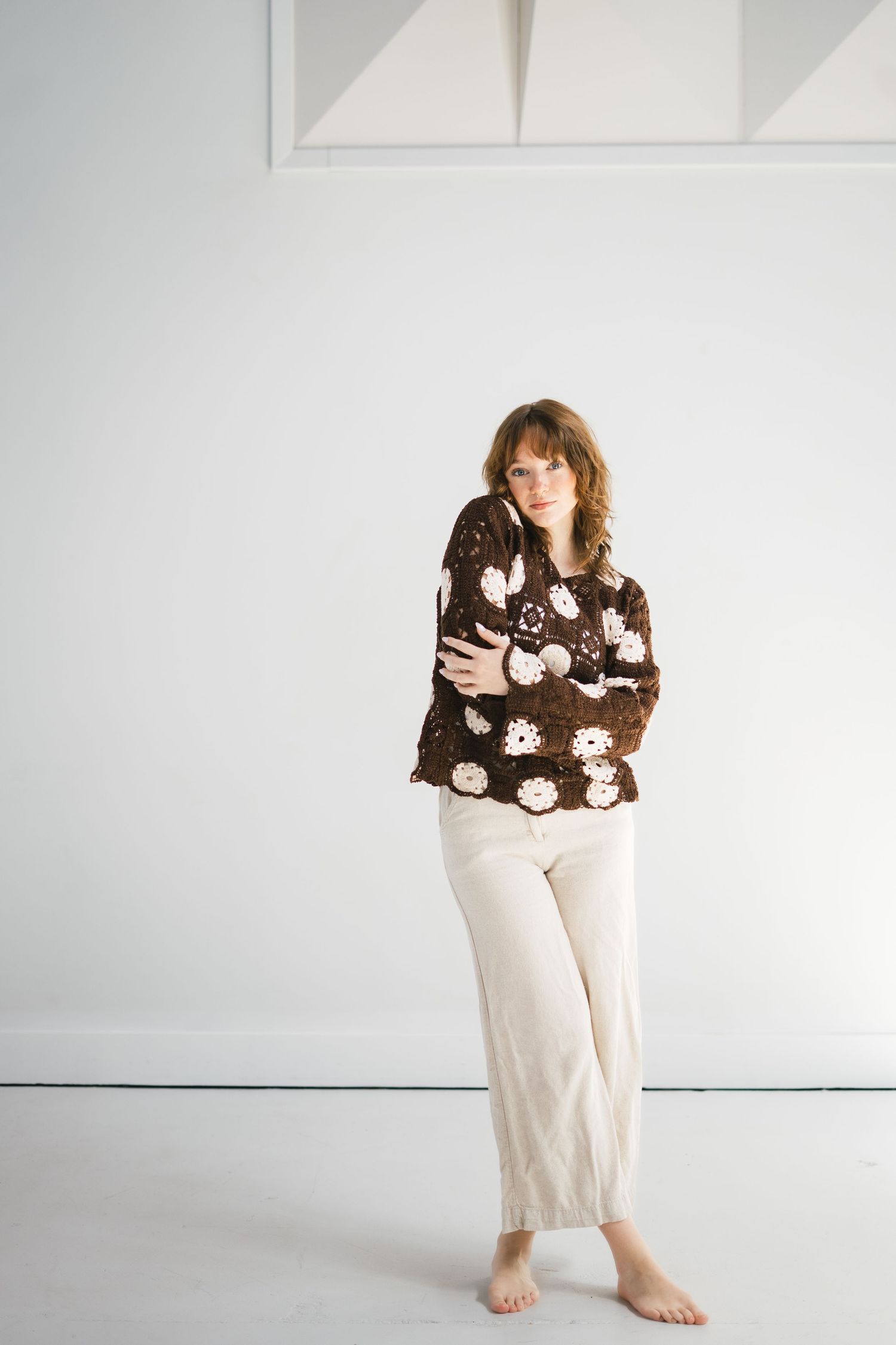 Woman wearing brown floral crochet sweater and cream wide-leg pants standing barefoot in white studio.