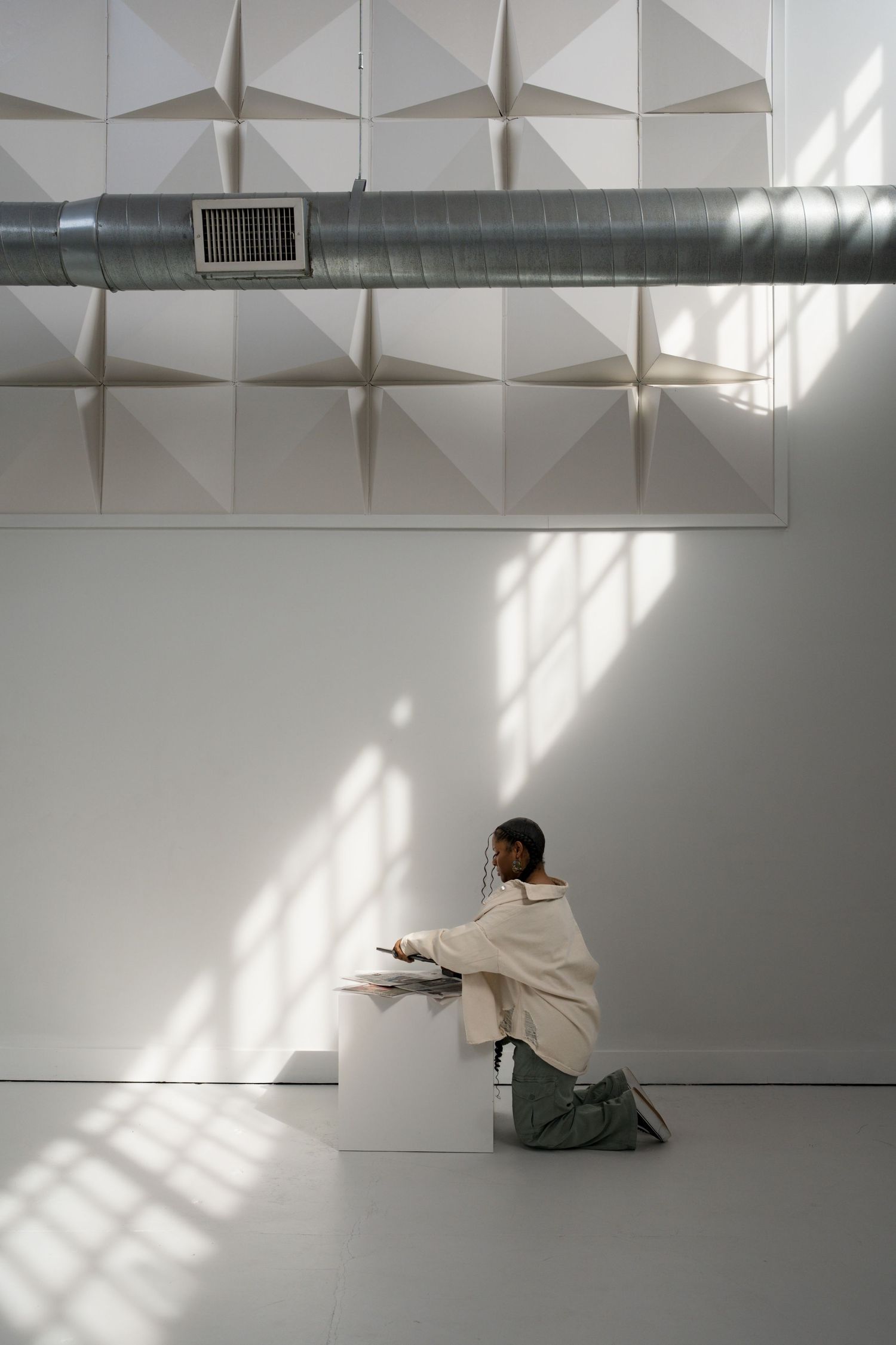 A Black woman in a white jacket kneels beside a white pedestal in a bright studio with geometric ceiling panels and exposed ductwork.