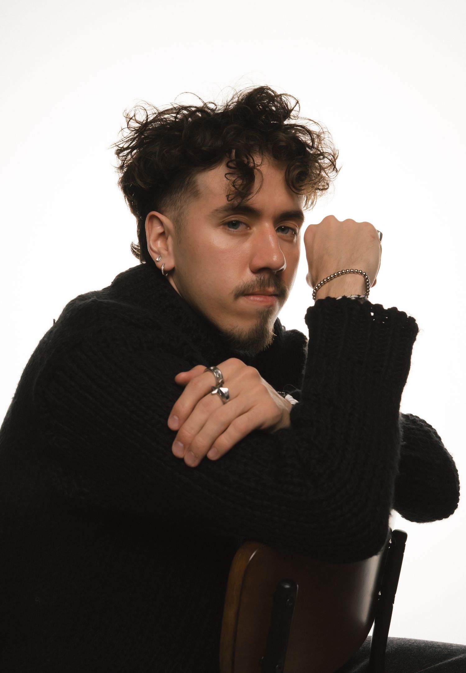 Young man in black knit sweater posing on chair with arms crossed, wearing silver rings and bracelet.