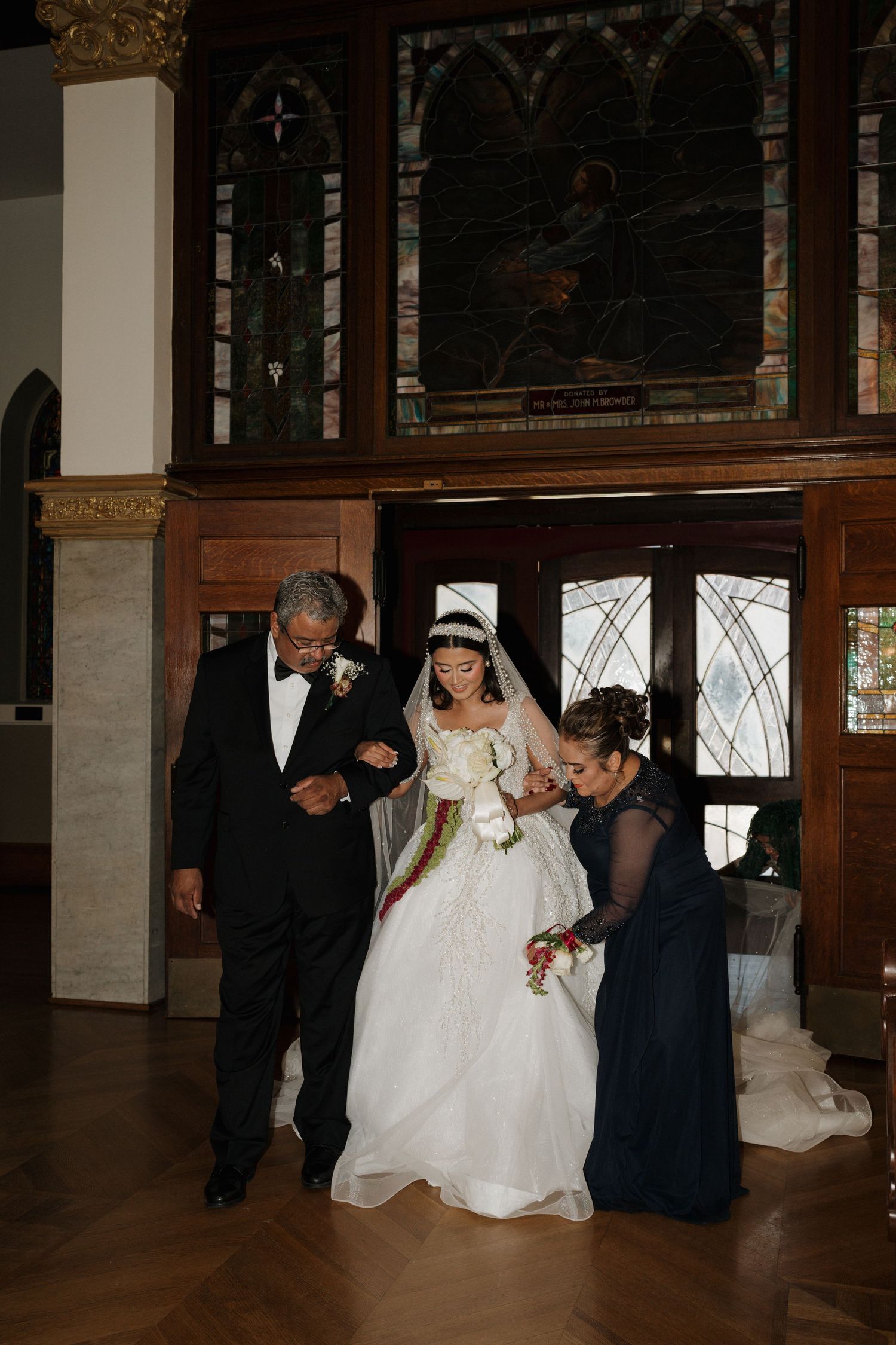 Bride in white ball gown being escorted by family members inside an ornate church entrance.