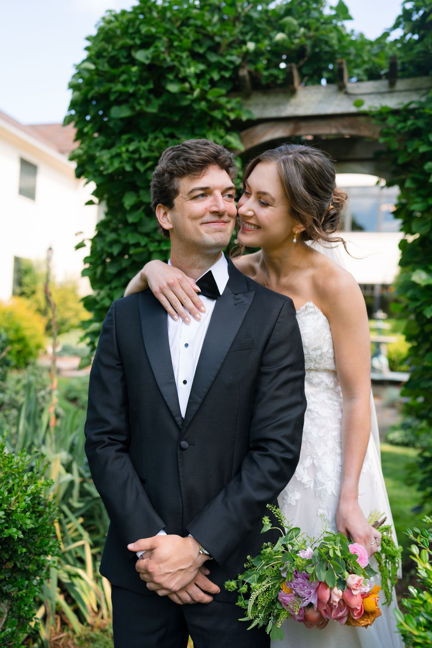 Bride embraces groom from behind under a vine-covered arch, both smiling in a garden setting.